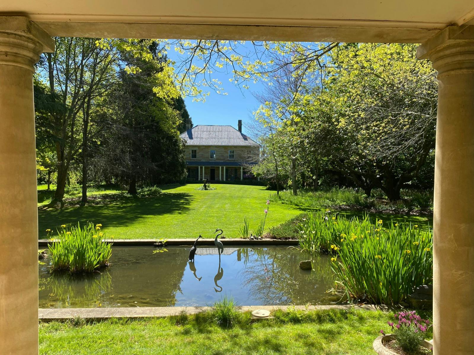 A grand mansion sits surrounded by trees and a luscious lawn leading down to a pond