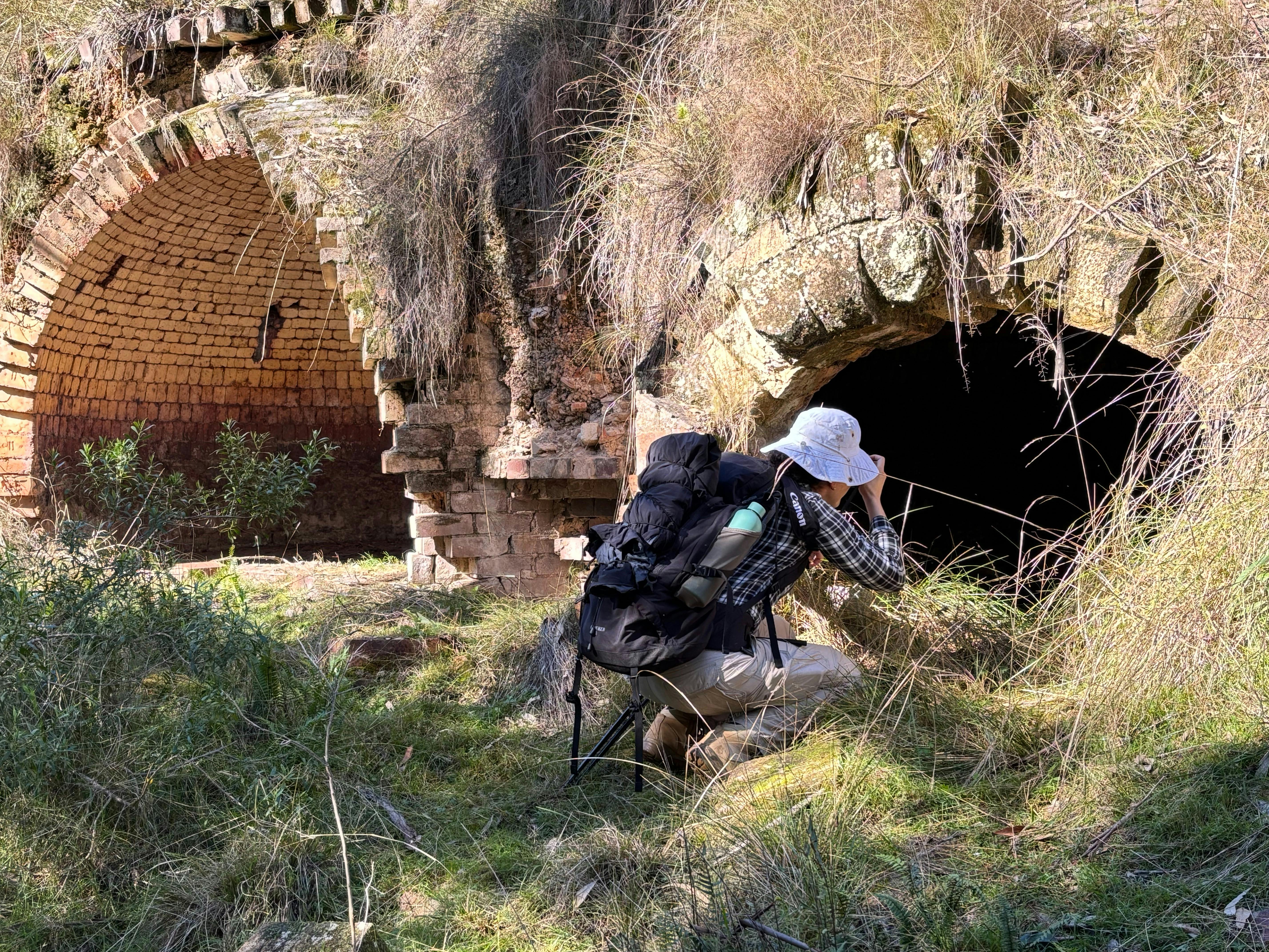 A woman wearing a checkered shirt crouching down in front of the ruins of a large brick coking oven.