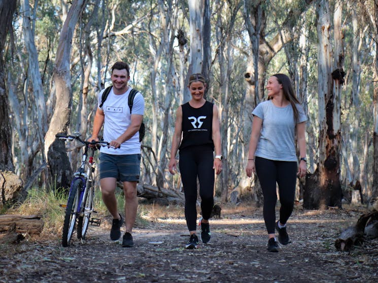 3 young adults hiking on a walking track in the Gunbower State Forest. One adult is pushing a bike