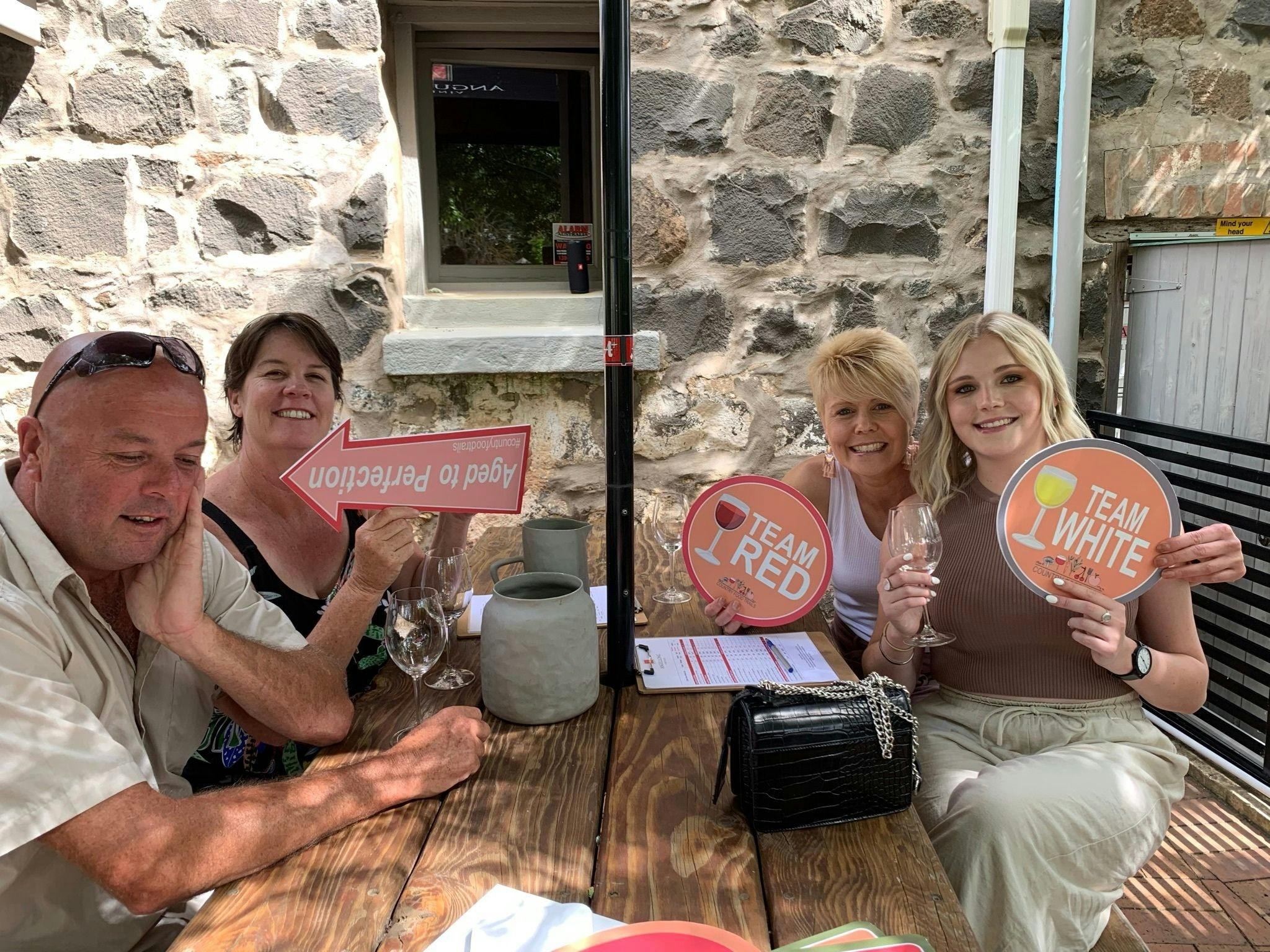 4 people sitting at table in courtyard tasting wine and holding signs in Millthorpe