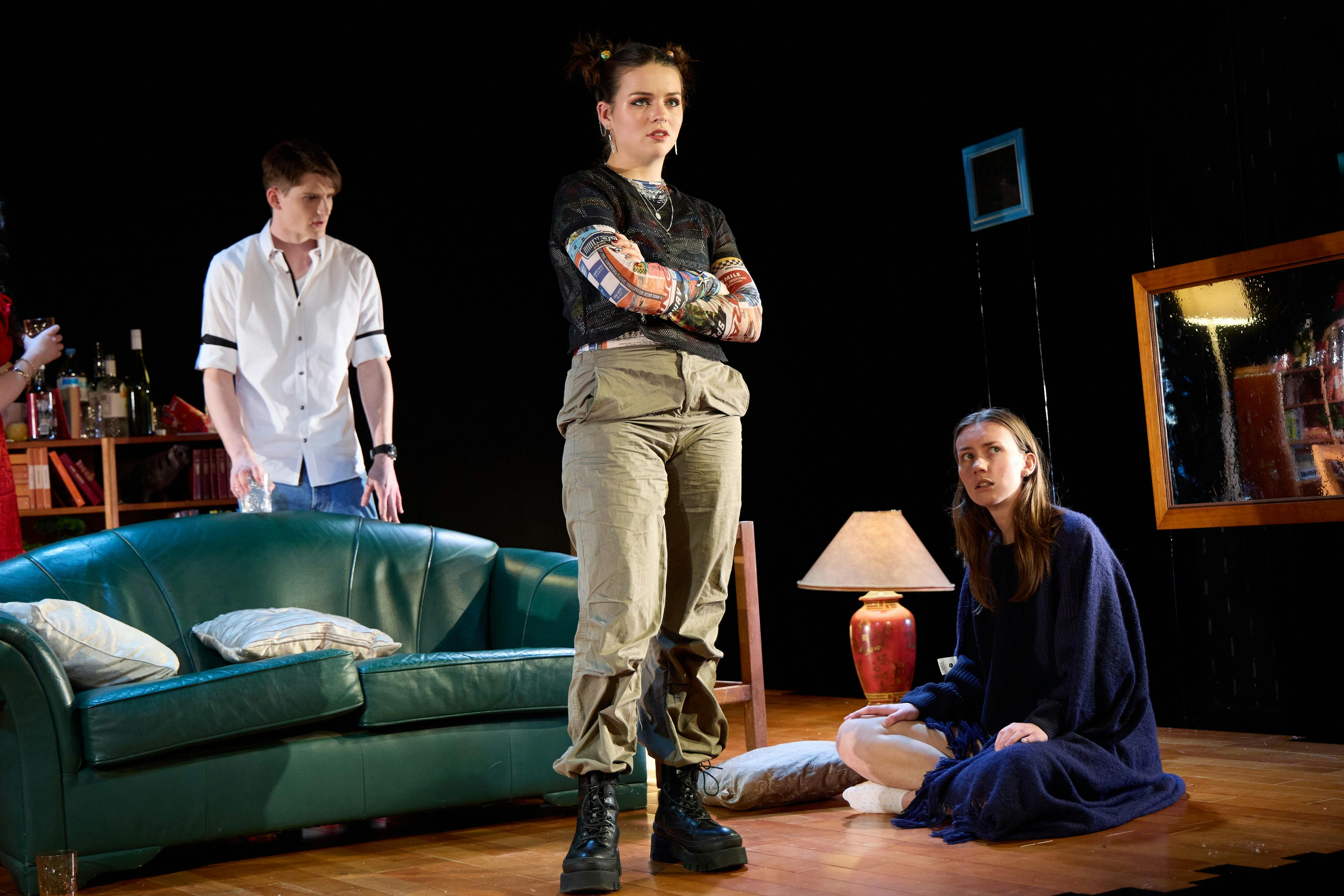 Three teenagers sit and stand in a stage set of a living room in a theatre production.