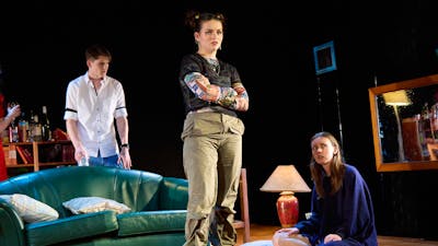 Three teenagers sit and stand in a stage set of a living room in a theatre production.