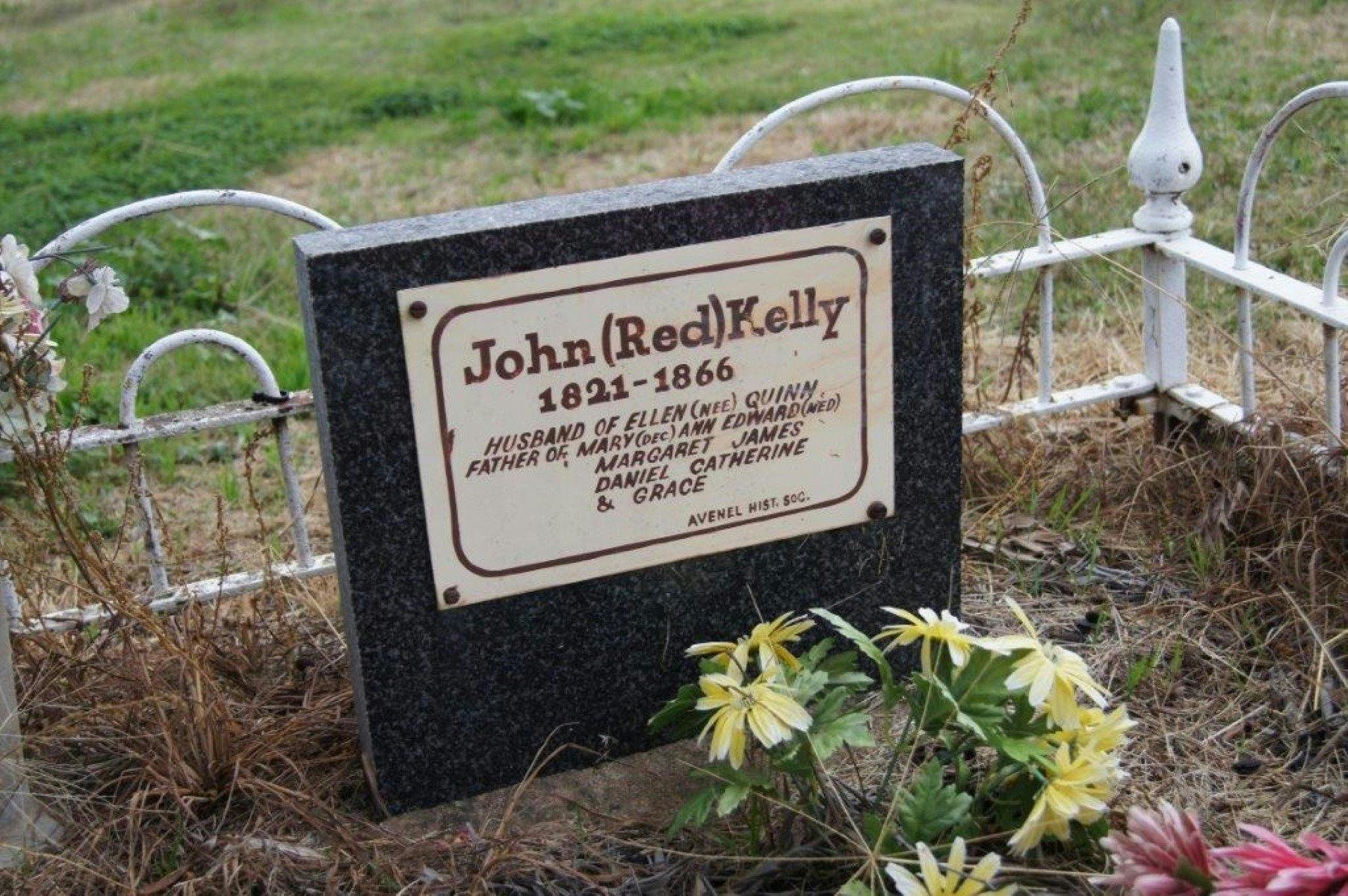a tombstone displaying the test John Red Kelly, surrounded by grass yellow flowers placed by it