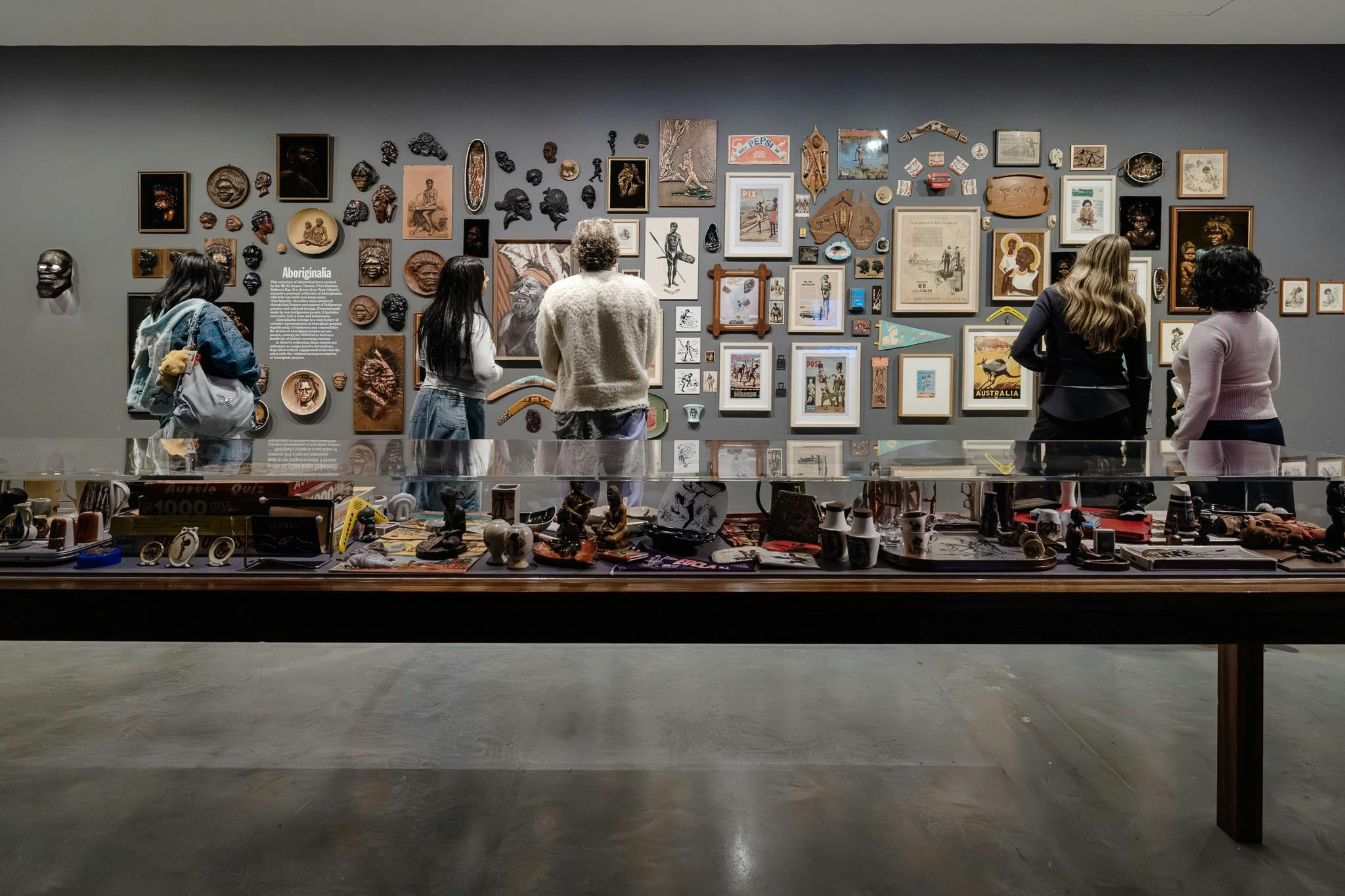 People view a wall of various artworks and objects, with a display case of objects in the foreground