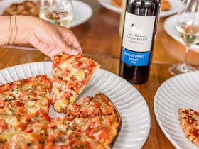hand grabbing pizza with bottles & glasses of wine