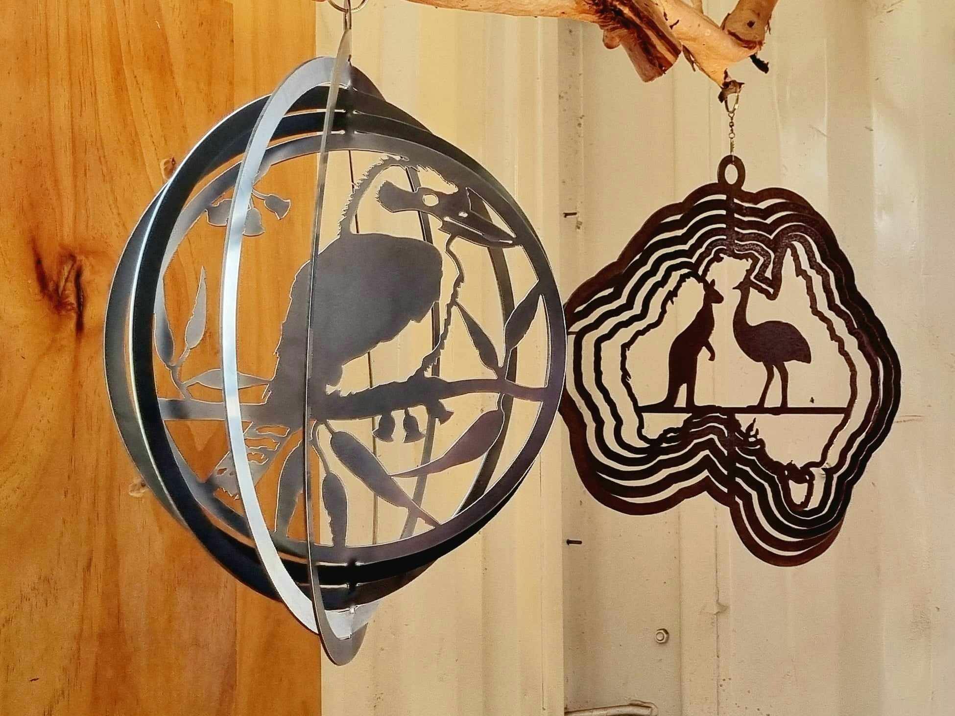 2 metal wind spinners, hanging from a branch against wood and white iron.