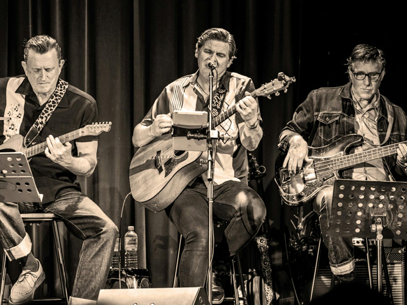 Three men seated on stage while playing guitars and singing