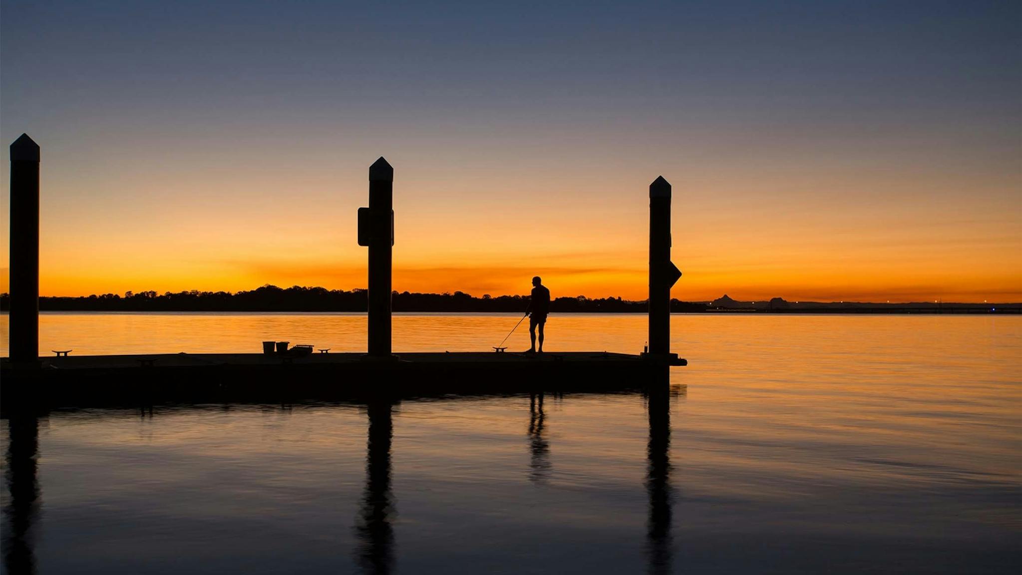The silhouette of a man fishing off a pier at Sunset, from Bongaree Beach Bribie Island