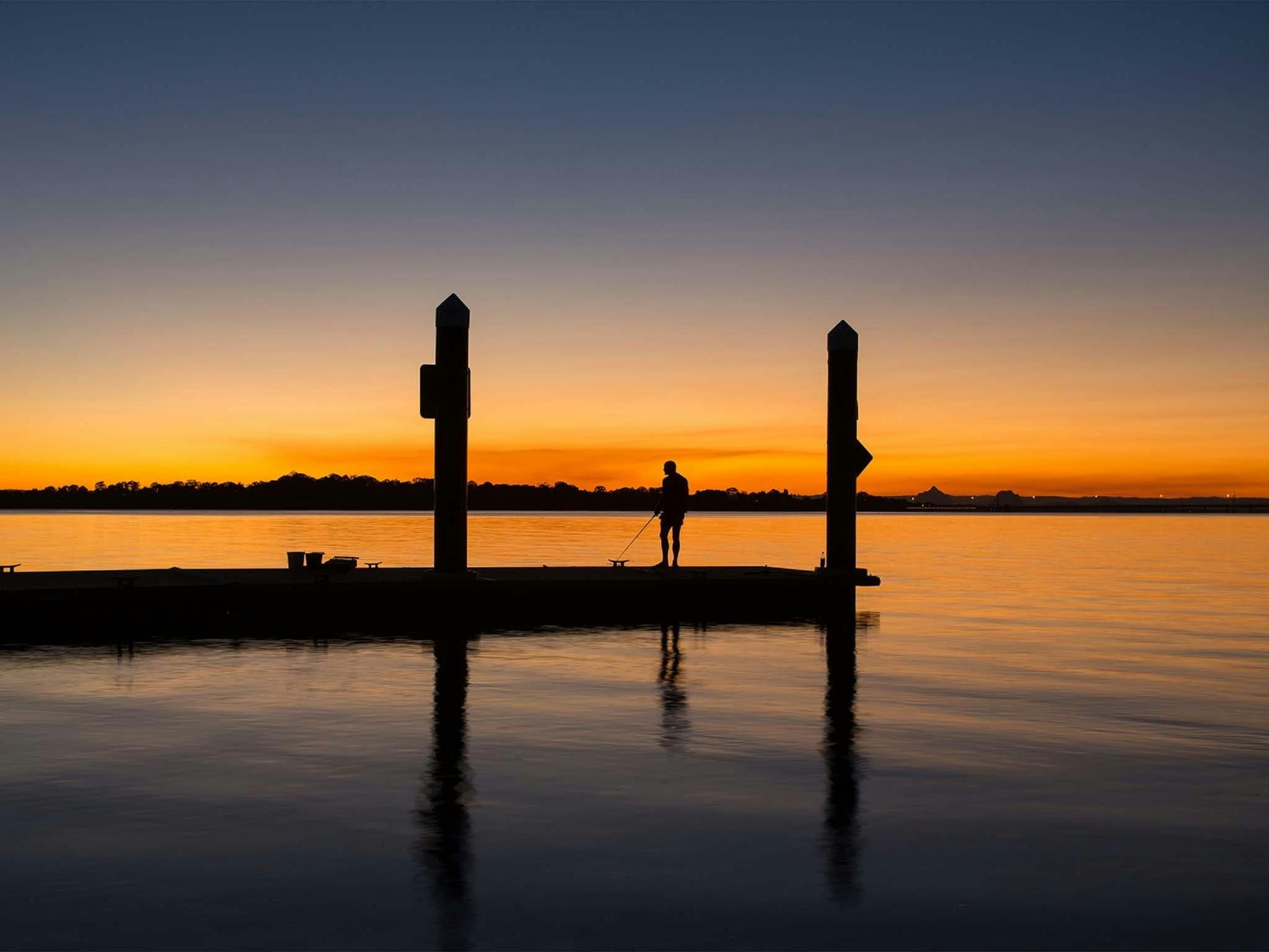 The silhouette of a man fishing off a pier at Sunset, from Bongaree Beach Bribie Island