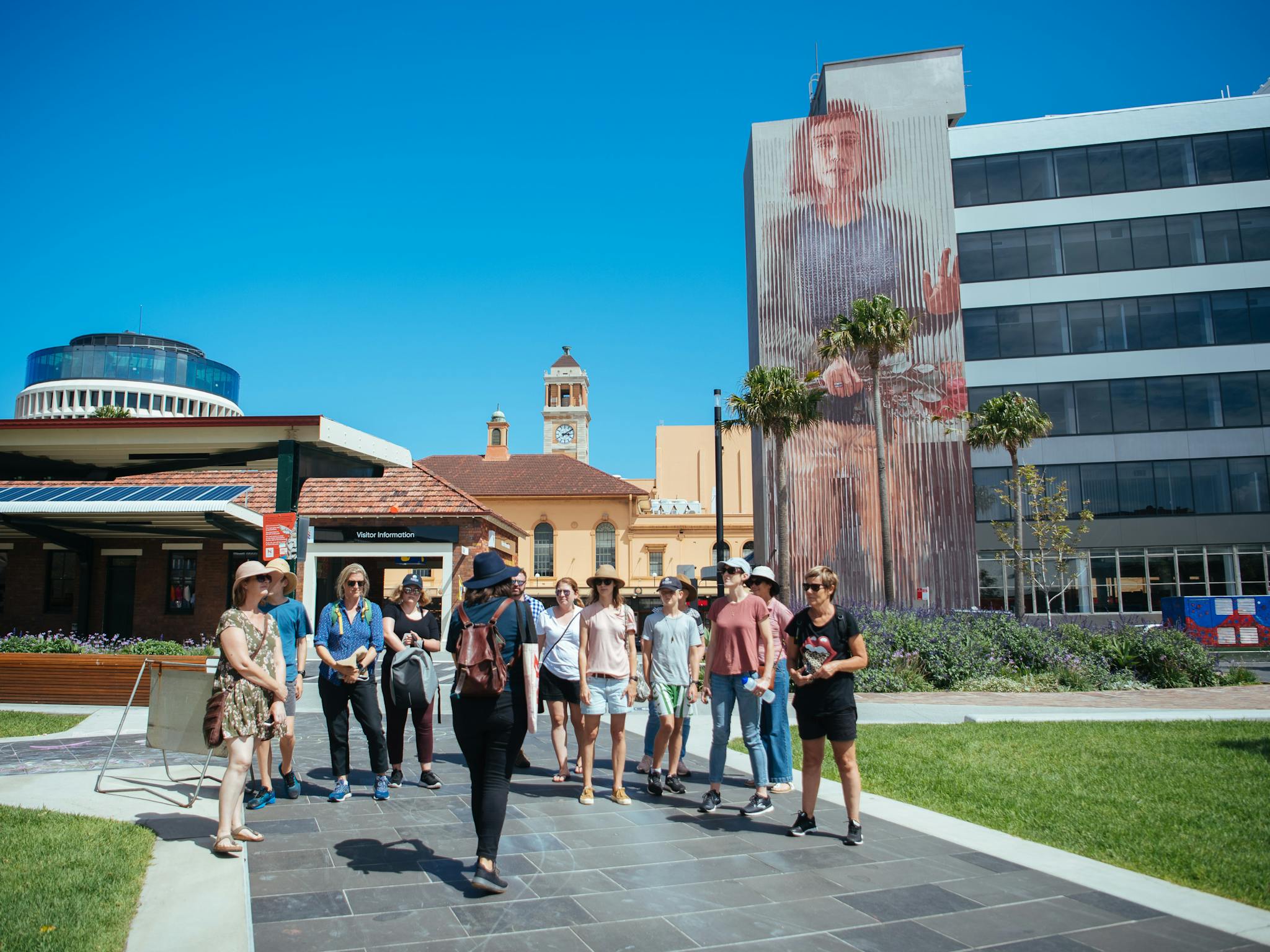 A group of people listen to a tour guide, standing in front of a large mural by Fintan Magee