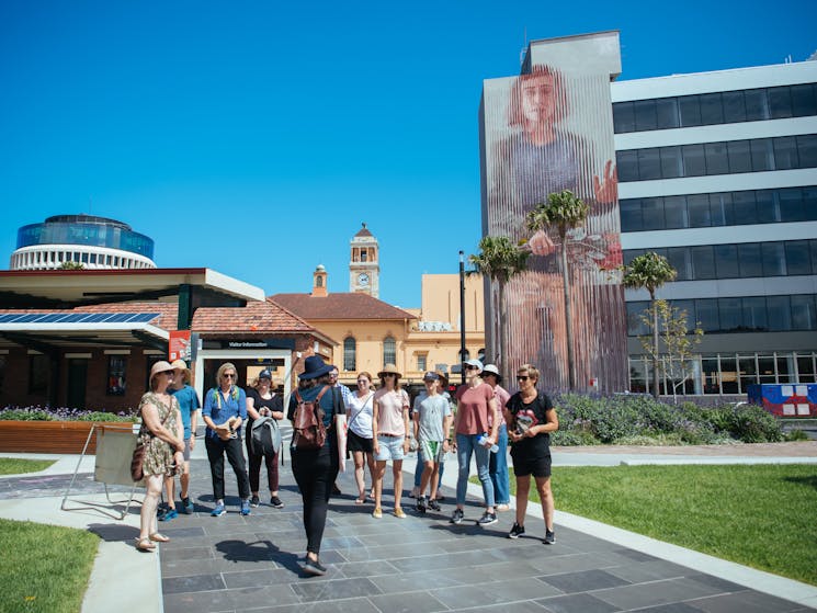 A group of people listen to a tour guide, standing in front of a large mural by Fintan Magee