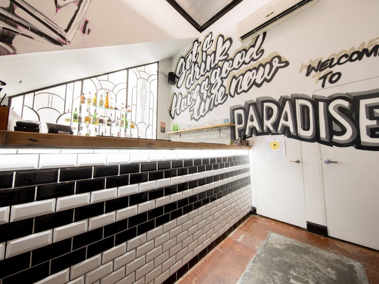 A bar with black and white subway tiles, a mural on the wall, and an art deco inspired back bar.