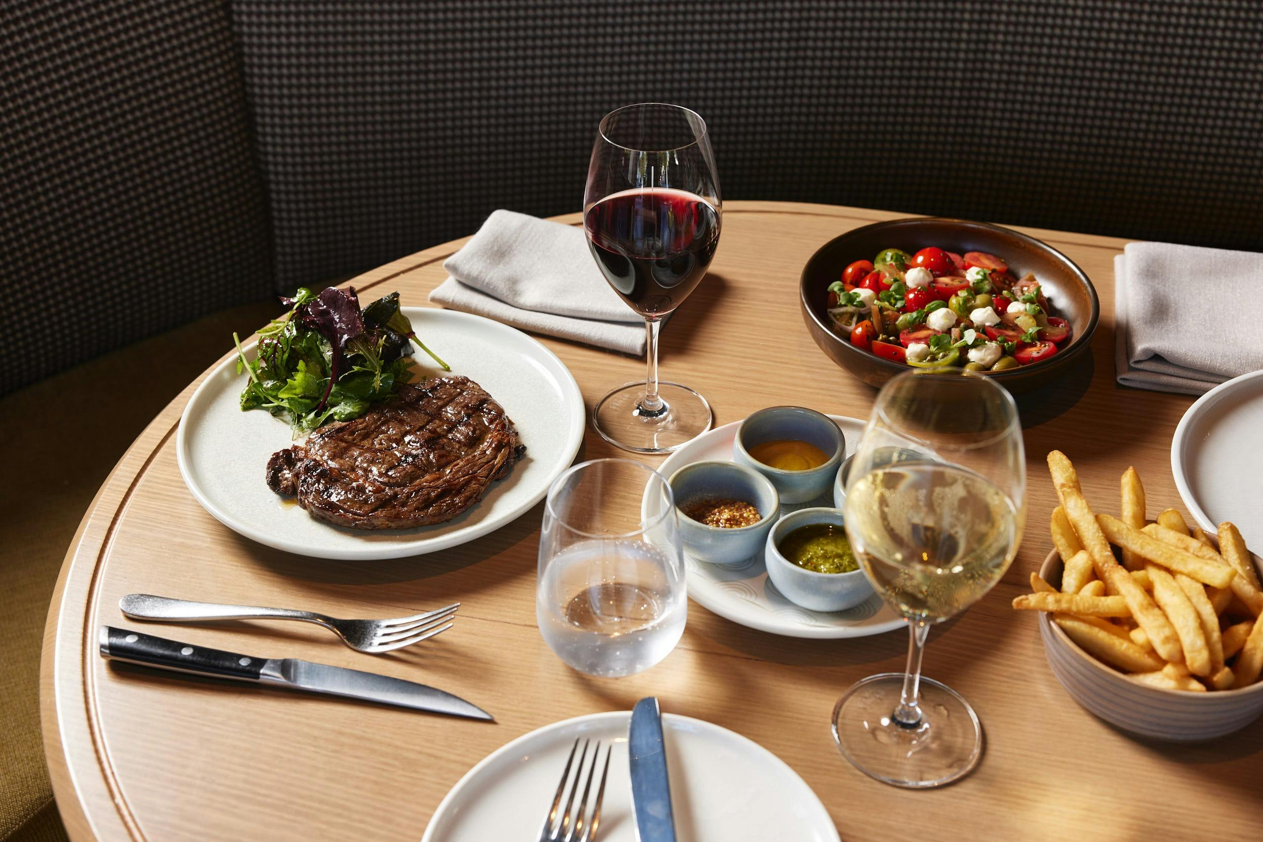 Steak with plate of sauces, bowl of fries, and glasses of wine on a table.