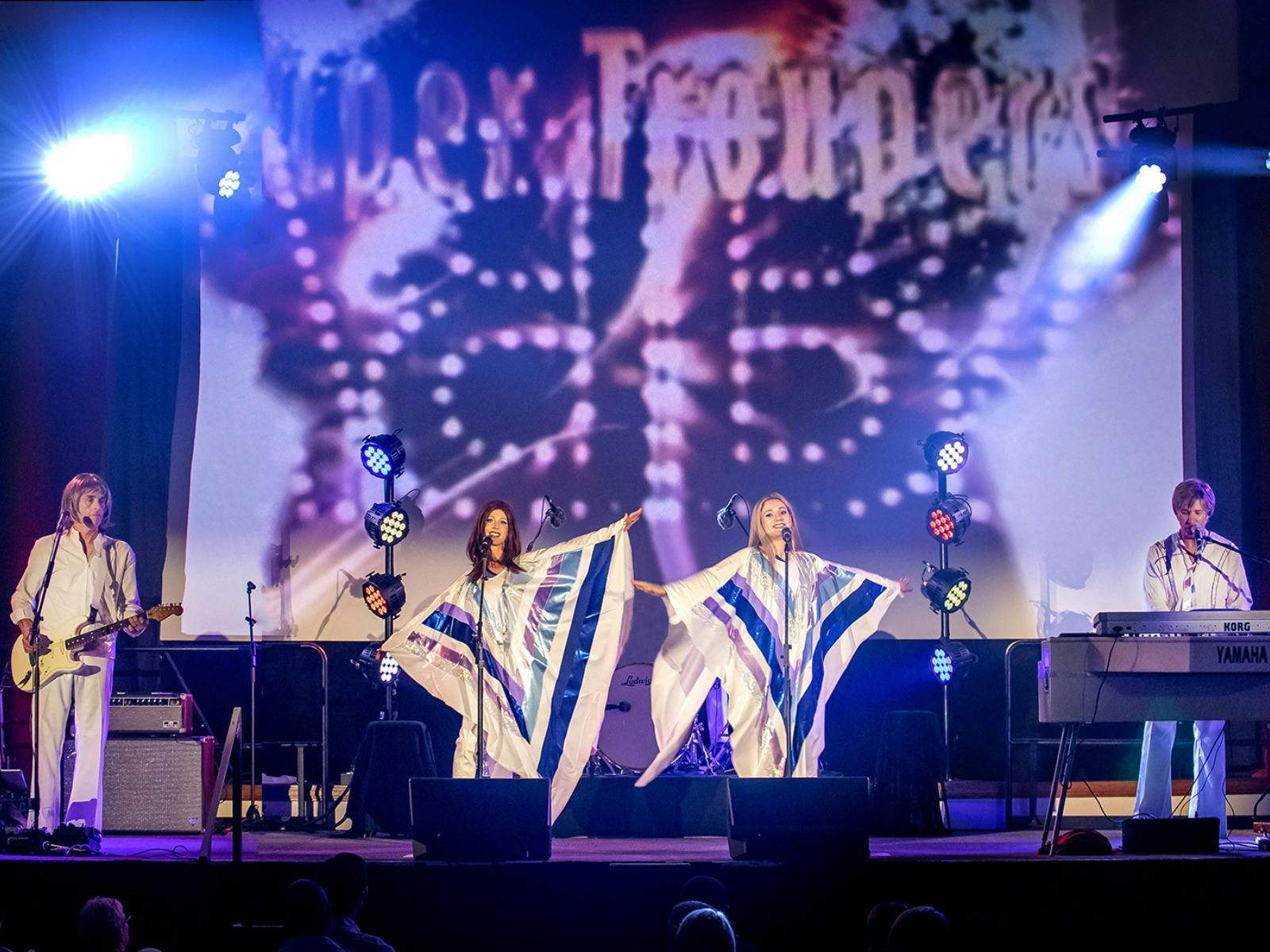 a group of 4 people performing in ABBA style costumes on a stage with lights and microphones
