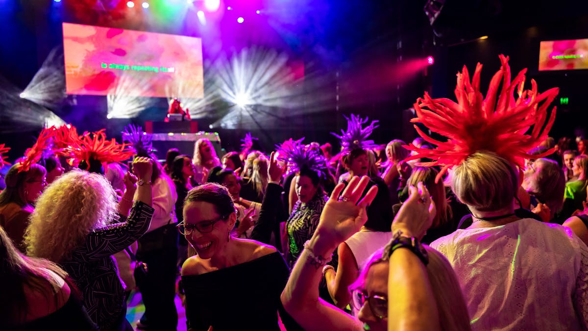 A crowd of ladies dance in front of the stage and bright stage lights, some wear feathered headbands