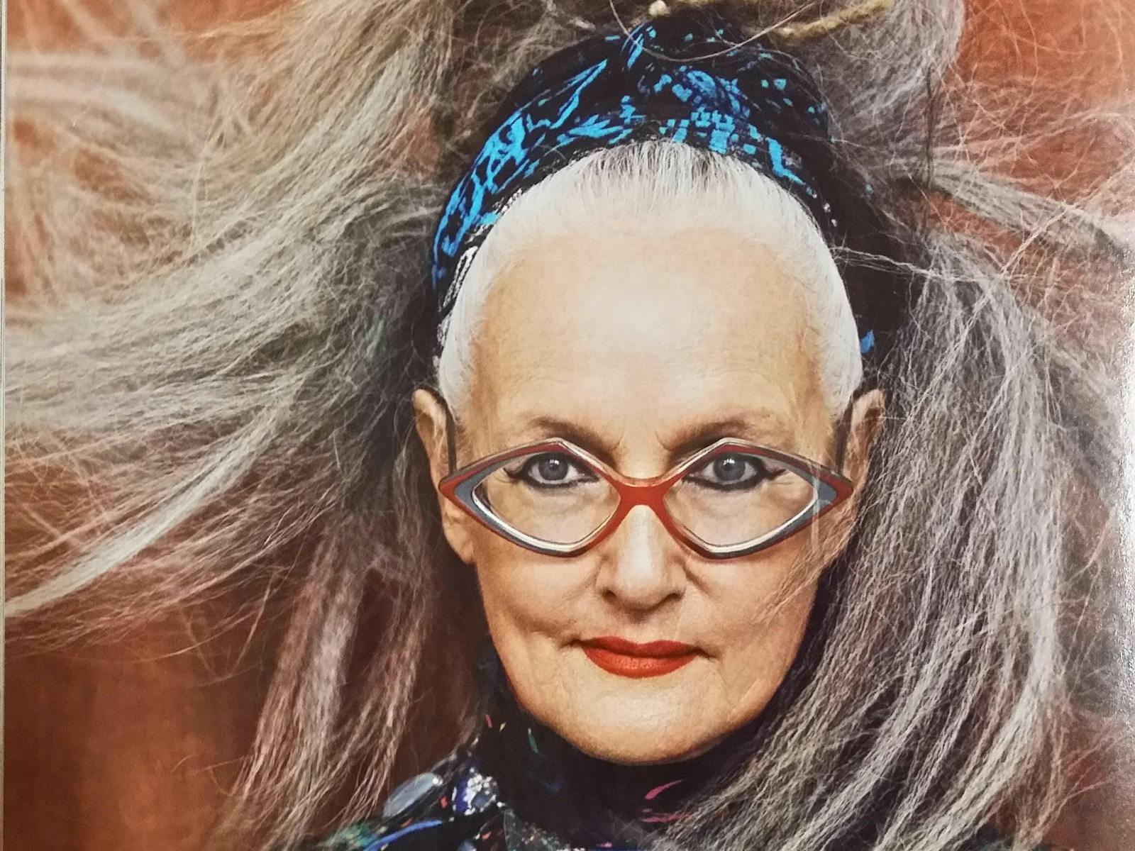 A woman with diamond-shaped classes smiles smartly. Her long grey ponytail sweeps around her.