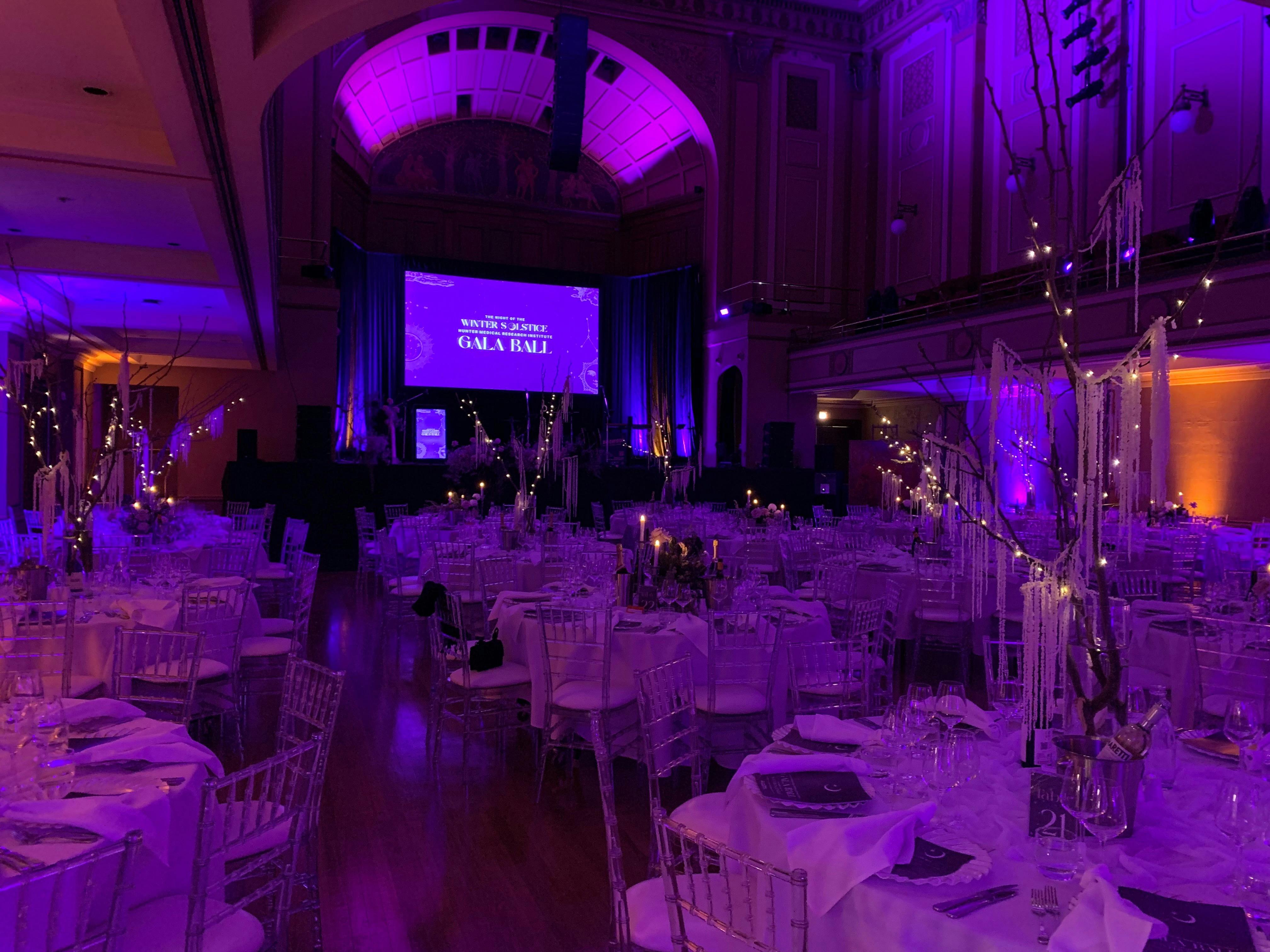 Concert Hall set for dinner with large round tables. The room is lit with purple mood lighting.