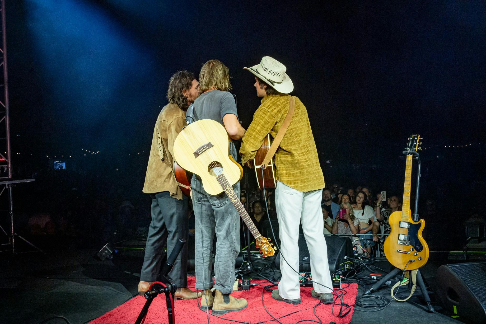 Three musicians singing around one microphone on a big stage. Their backs are to the camera.