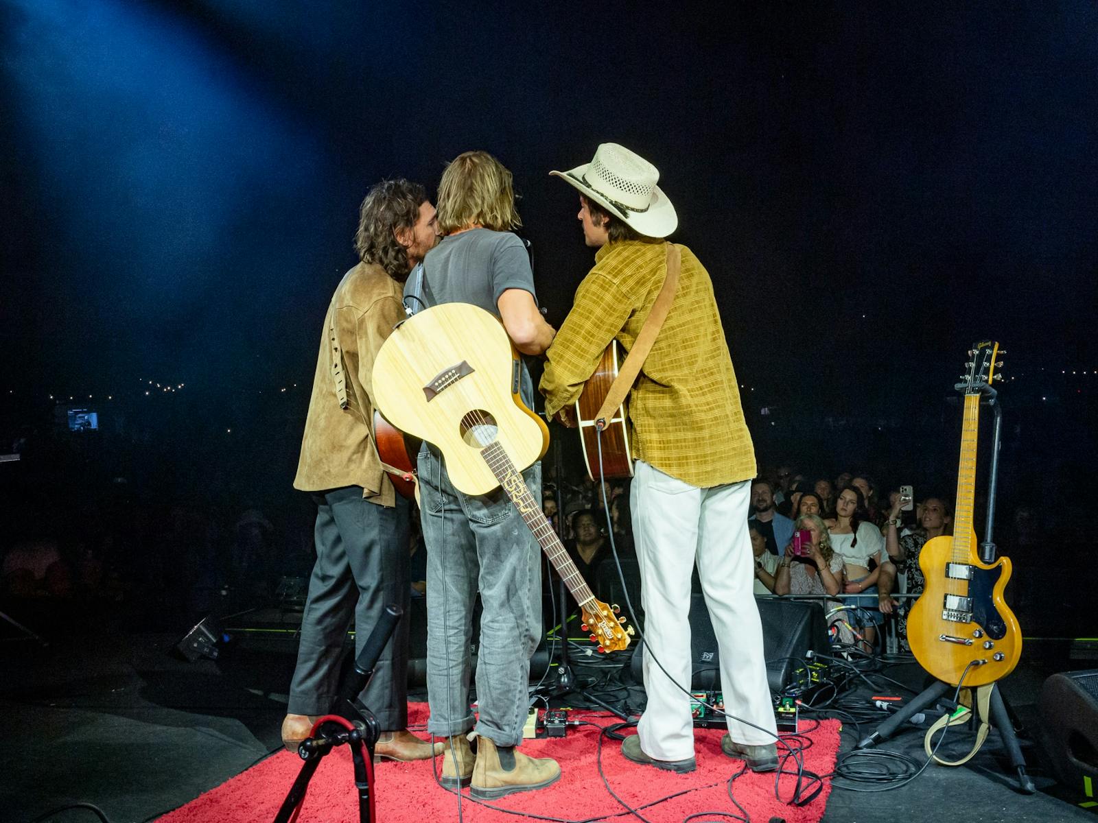 Three musicians singing around one microphone on a big stage. Their backs are to the camera.