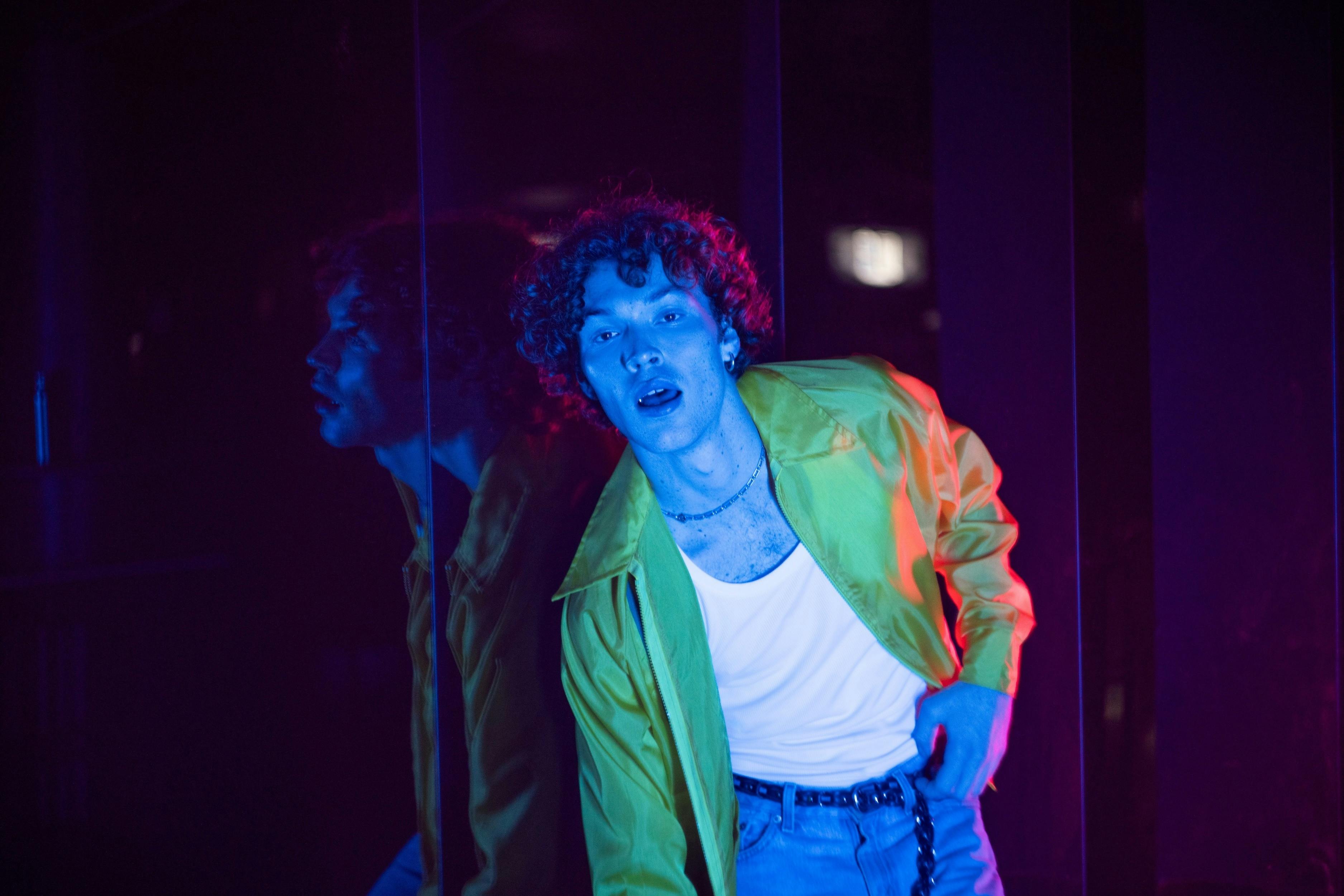Man wearing white singlet, green jacket and jeans poses with blue light washing over him.