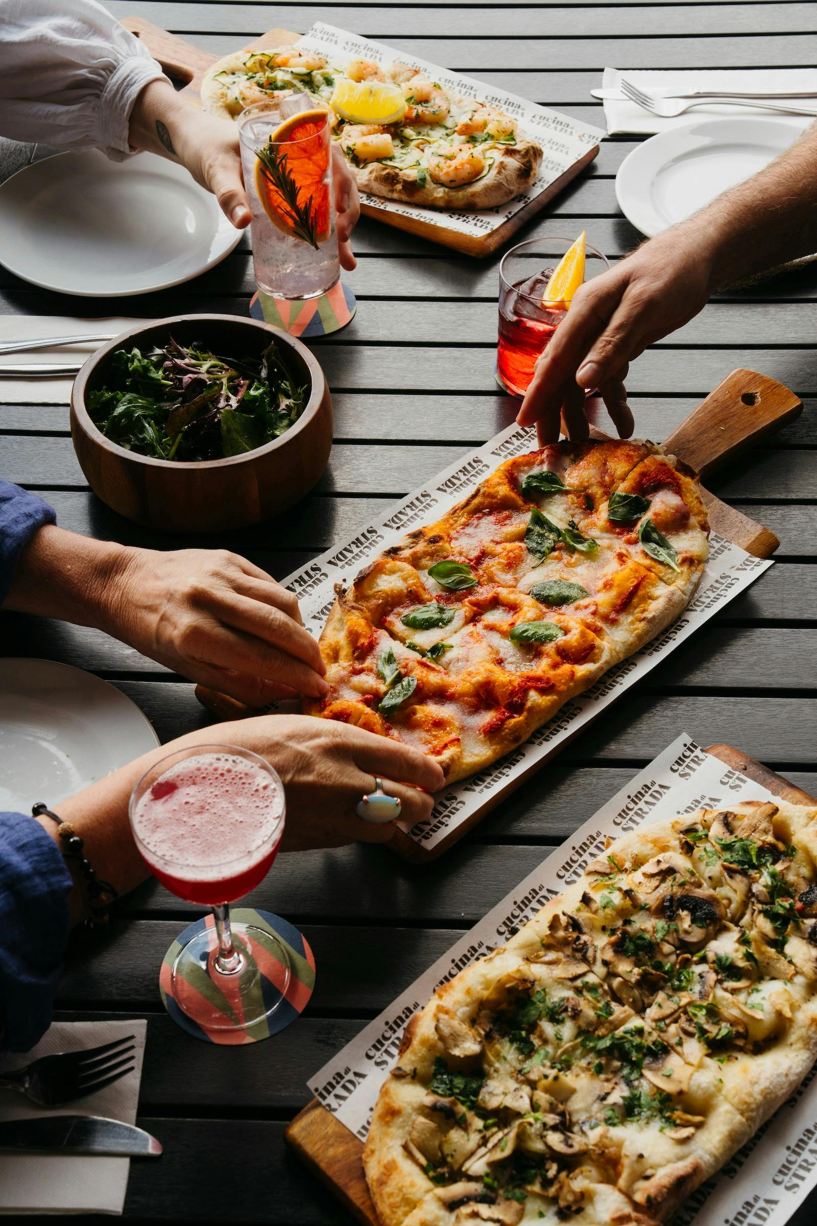 Pinsa and bowl of salad leave being reached for with a pink cocktail in a glass