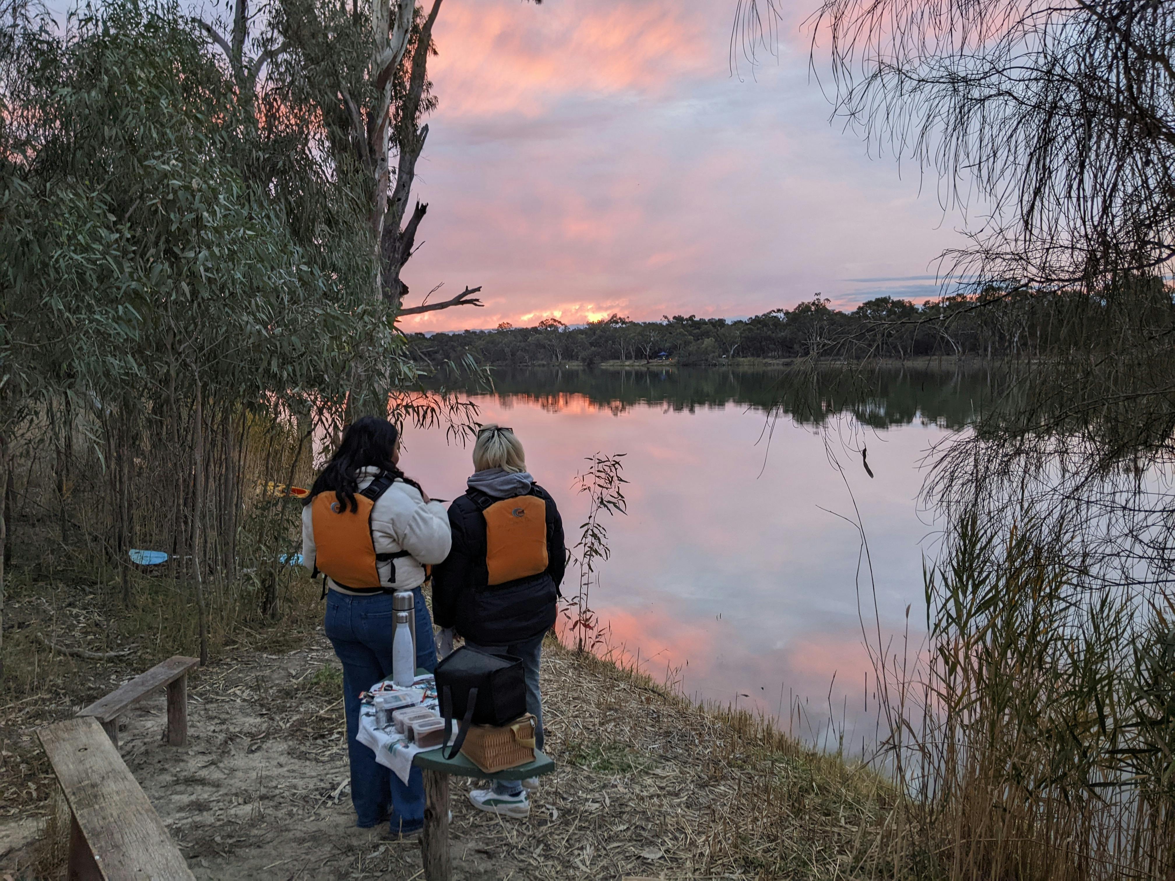2 people wearing life jackets stand on the water's edge, viewing the sunset reflected on the water.