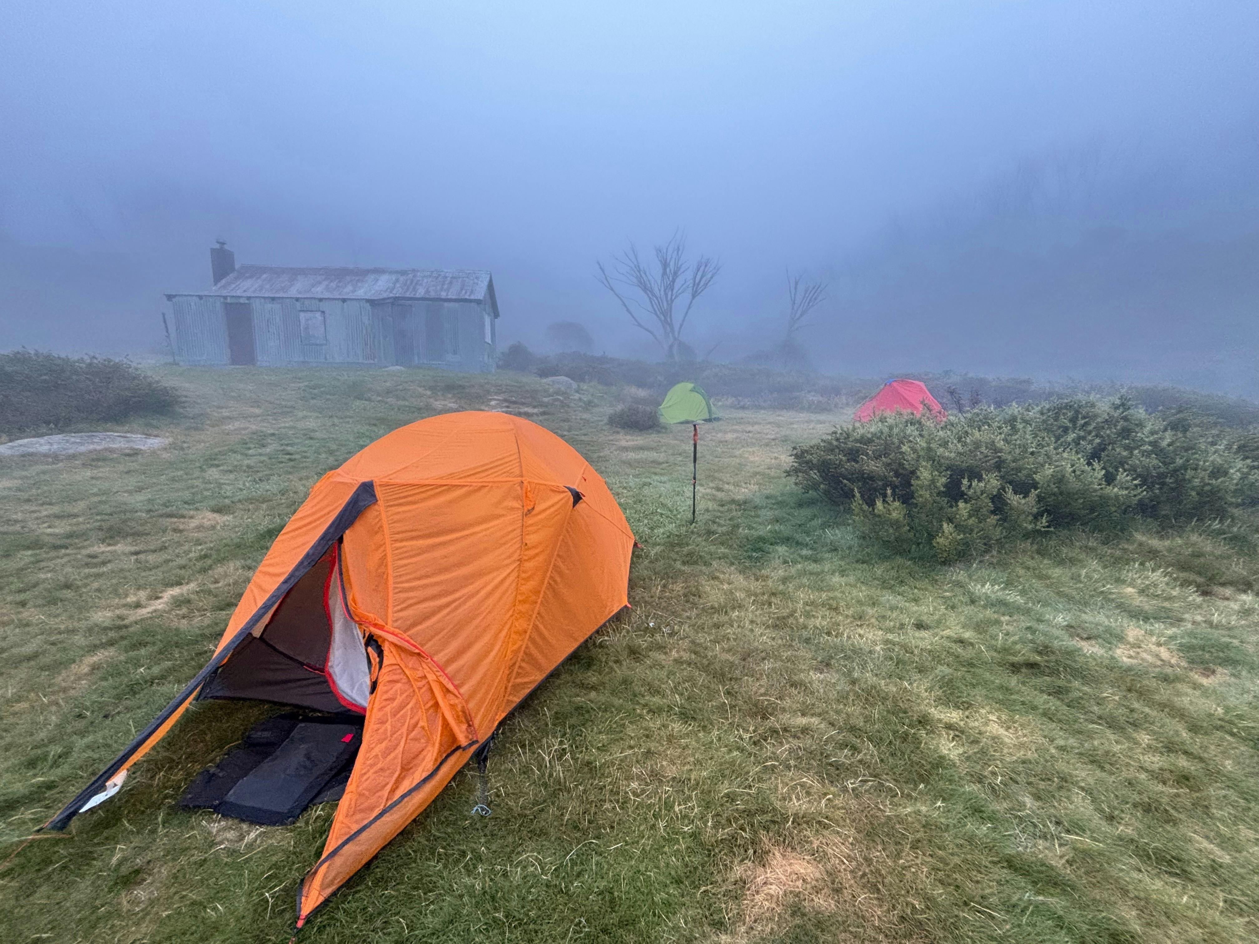 A metal galvanized hut and bright orange tent shrouded in early morning fog.
