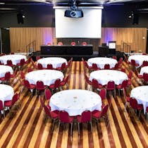 Round tables & chairs, projection screen, wooden floorboards, straight table for presenters at front
