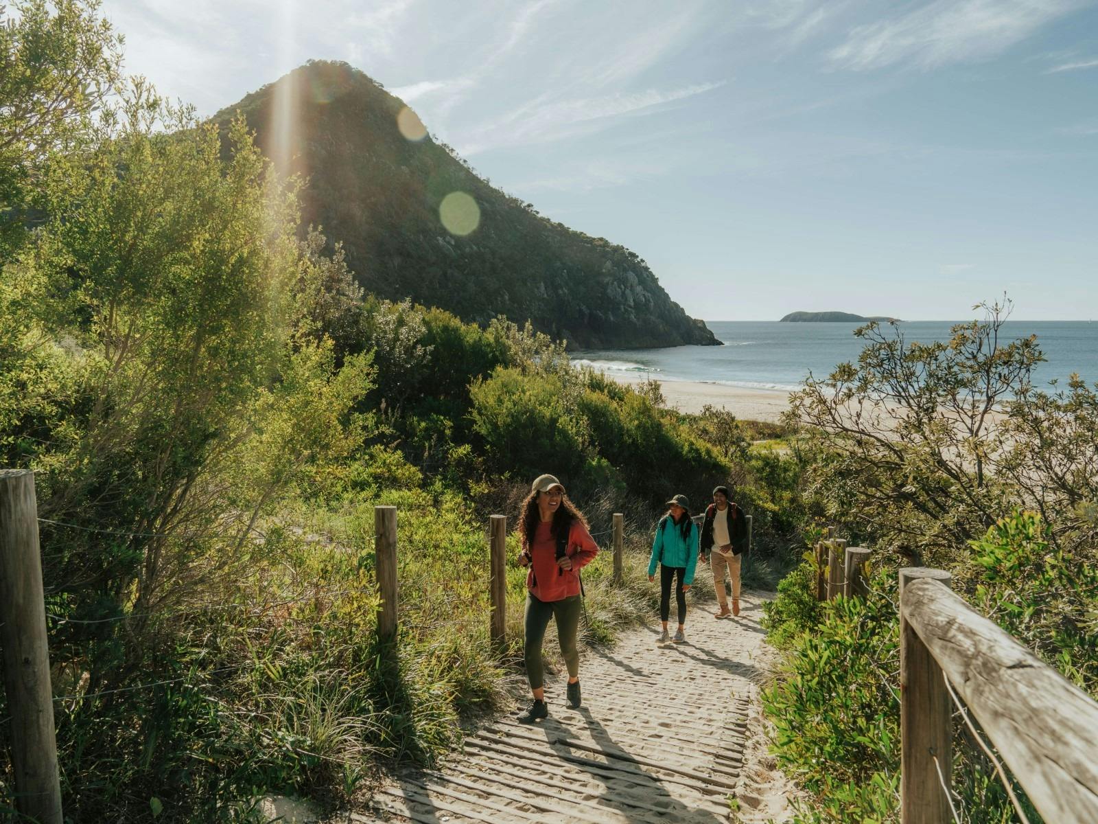 Three people walking on a path from Zenith Beach, with ocean and Tomaree Summit in background