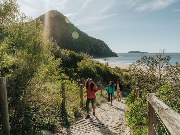 Three people walking on a path from Zenith Beach, with ocean and Tomaree Summit in background