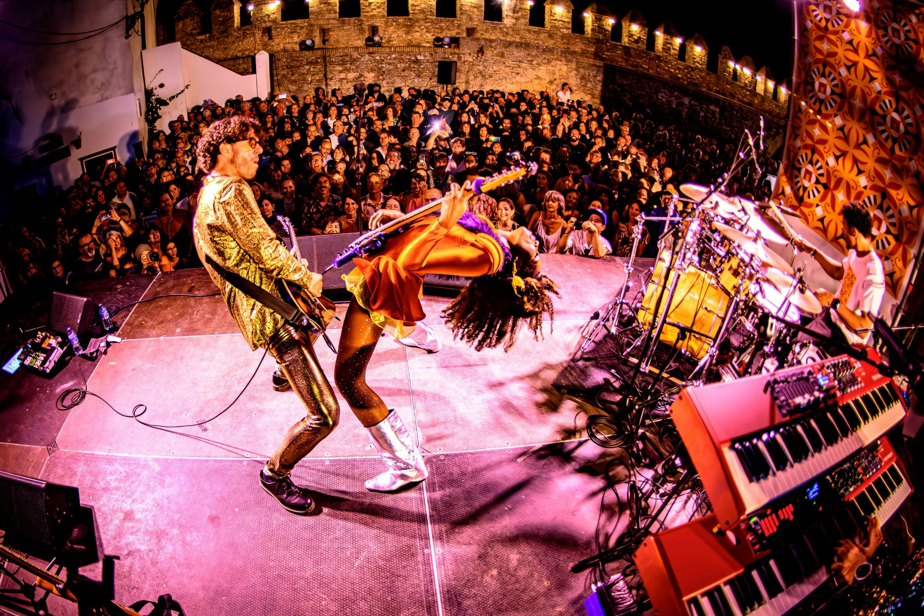 Woman playing bass guitar with man in front of large crowd.