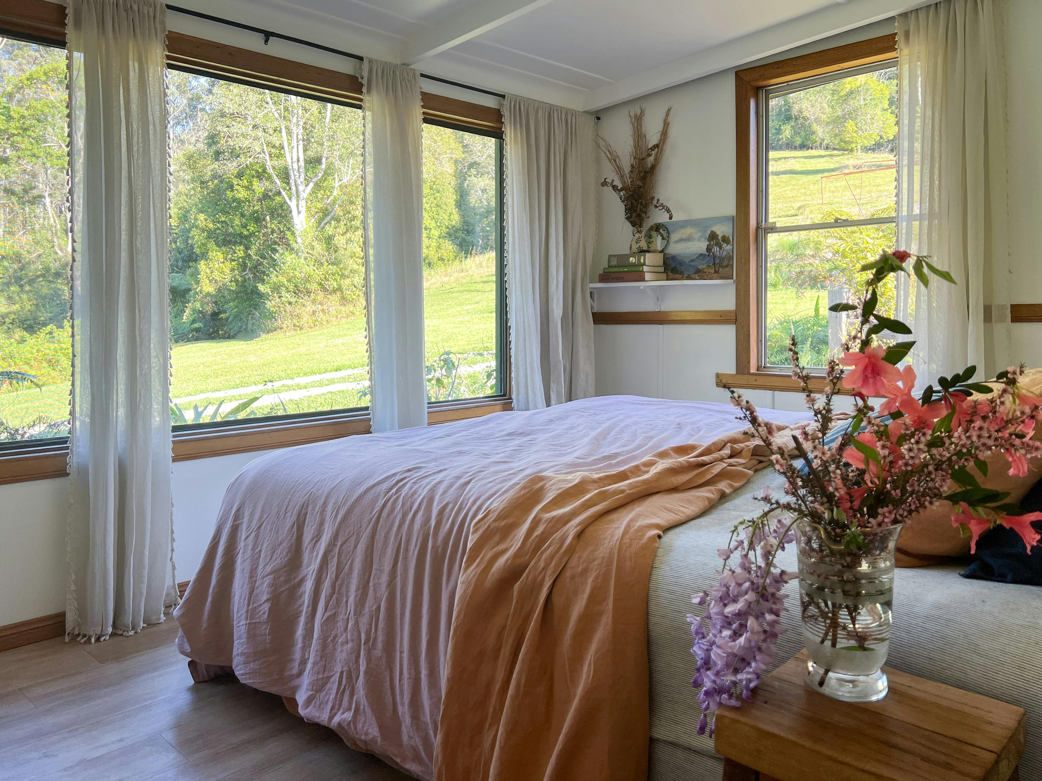 A dreamy bedroom with boho curtains, ochre bedding and a large window with a forest view