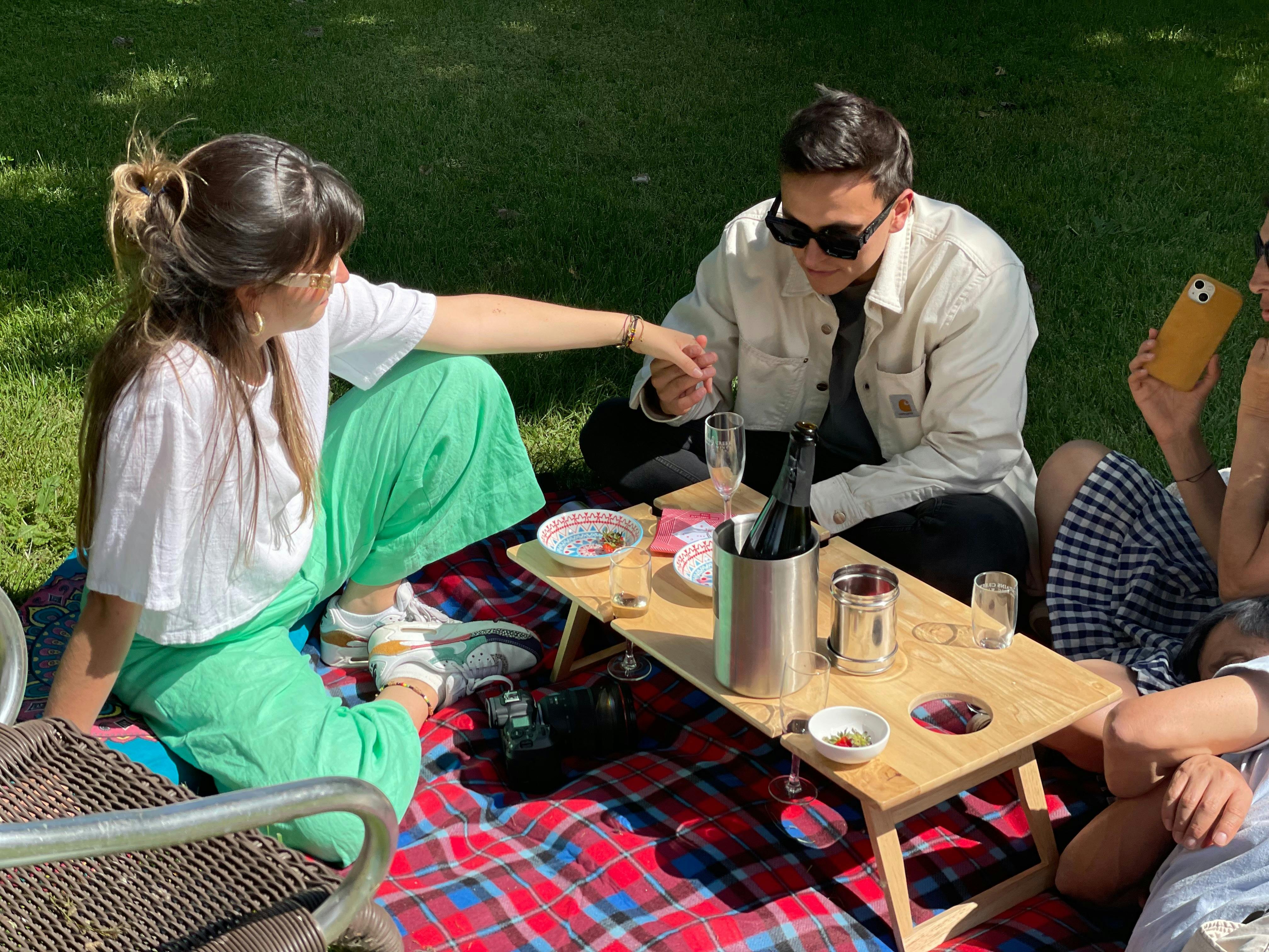 a man and woman in their late 20s hold hands while sitting on a picnic rung and cushions.  Wine.