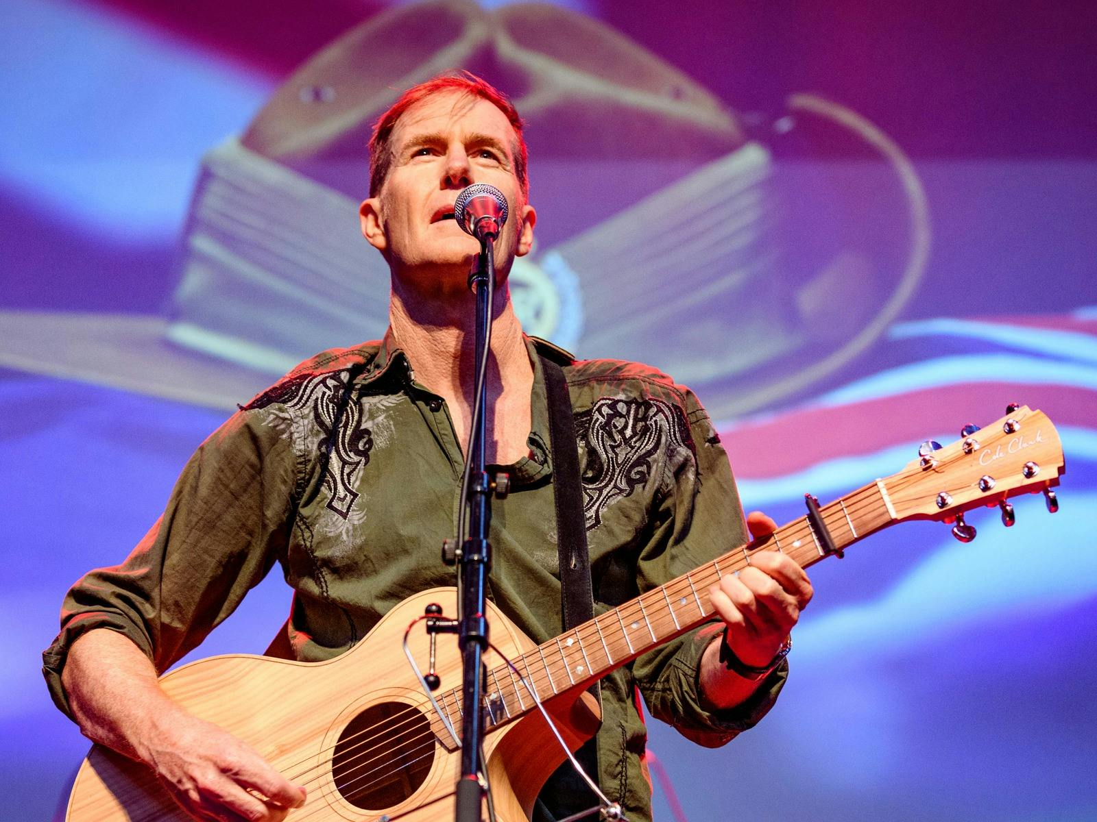 A white man in a khaki shirt standing at a mic and playing a guitar