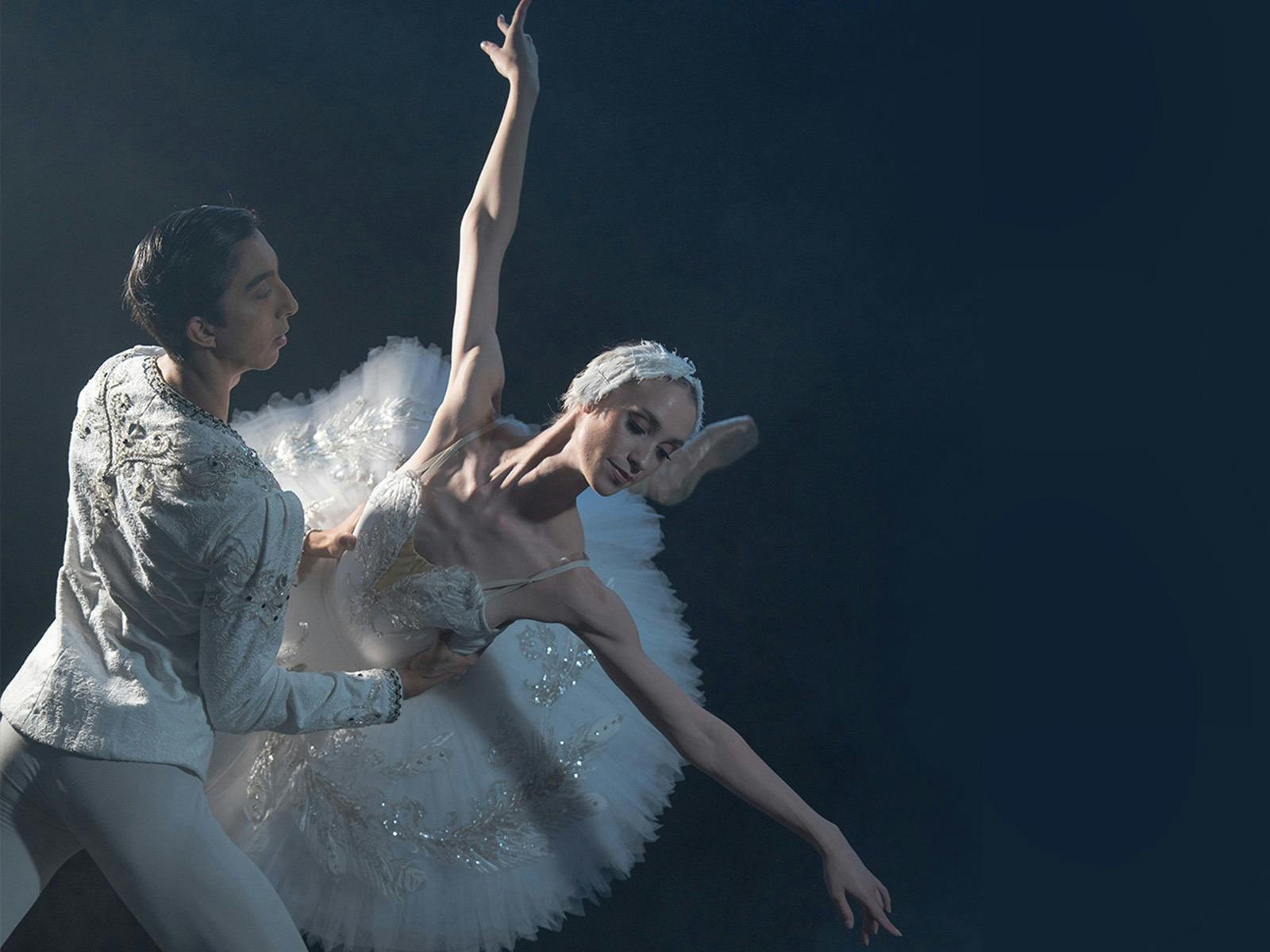 Two ballet dancers in white costumes perform a dramatic pose from Swan Lake on a dark stage