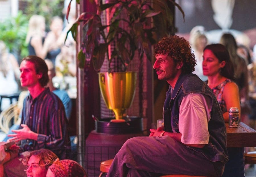 People seated indoors watching live entertainment under pink lighting, drinks in hand.