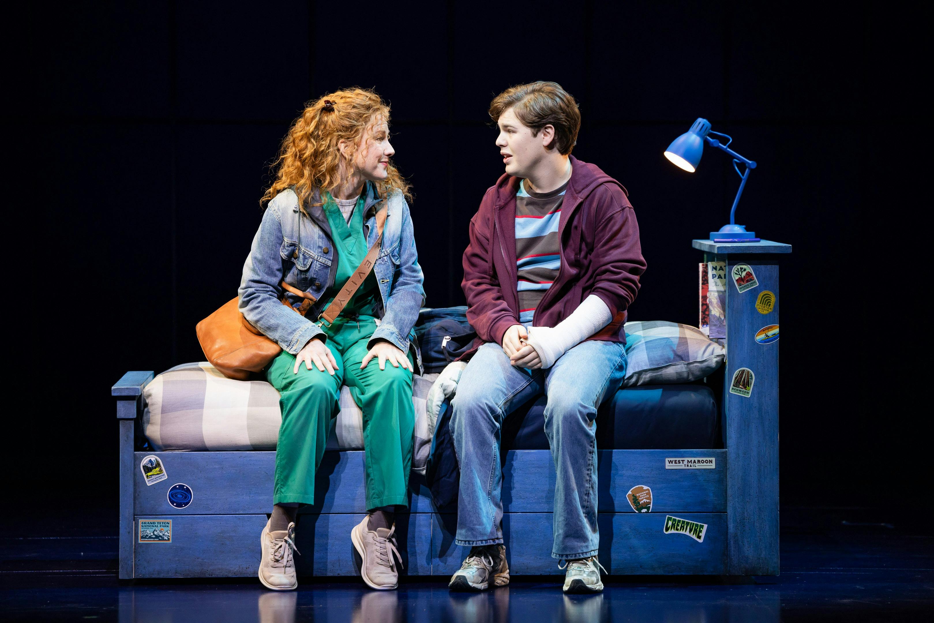 A mother and a son sit on a bed in conversation.