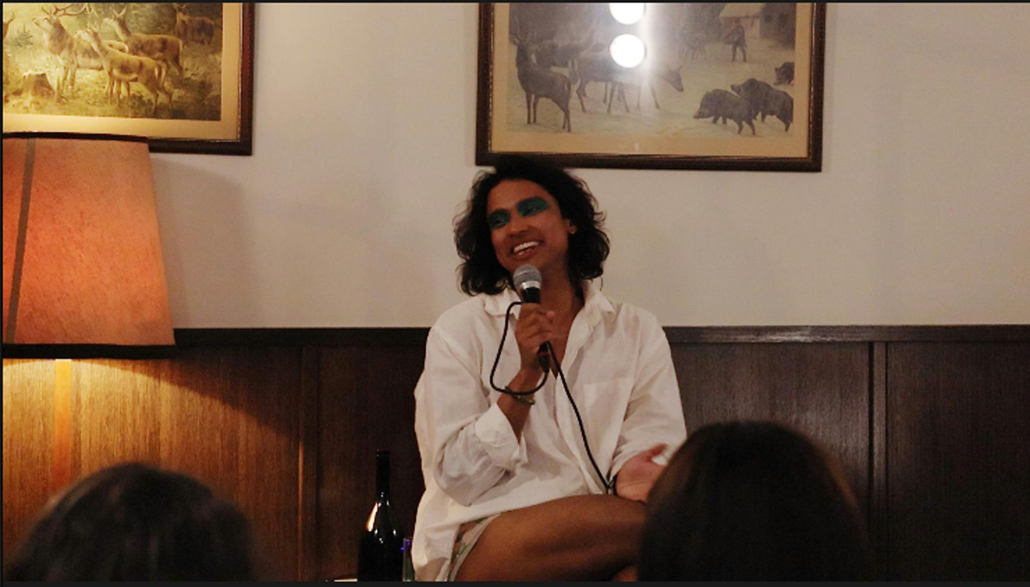 Neptune sits on a chair, holding a microphone, in a white shirt. He is a brown person with dark hair