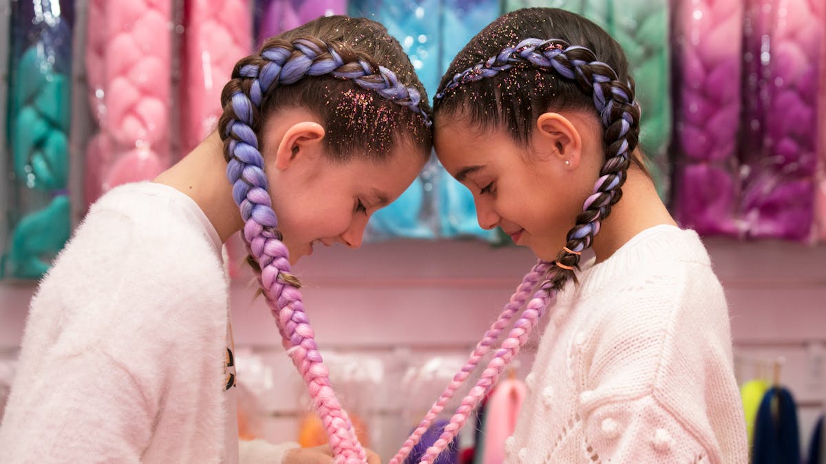 two girls with their heads together and their hair braids are forming a love heart shape