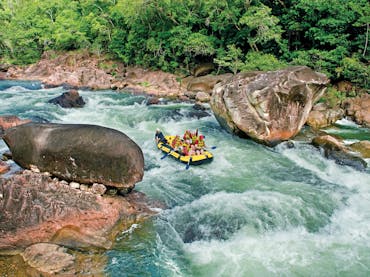 Wide shot of a rafting boat surrounded by blue water, lush rainforest and large boulders in Tully Wide shot of a rafting boat surrounded by blue water, lush rainforest and large boulders in Tully