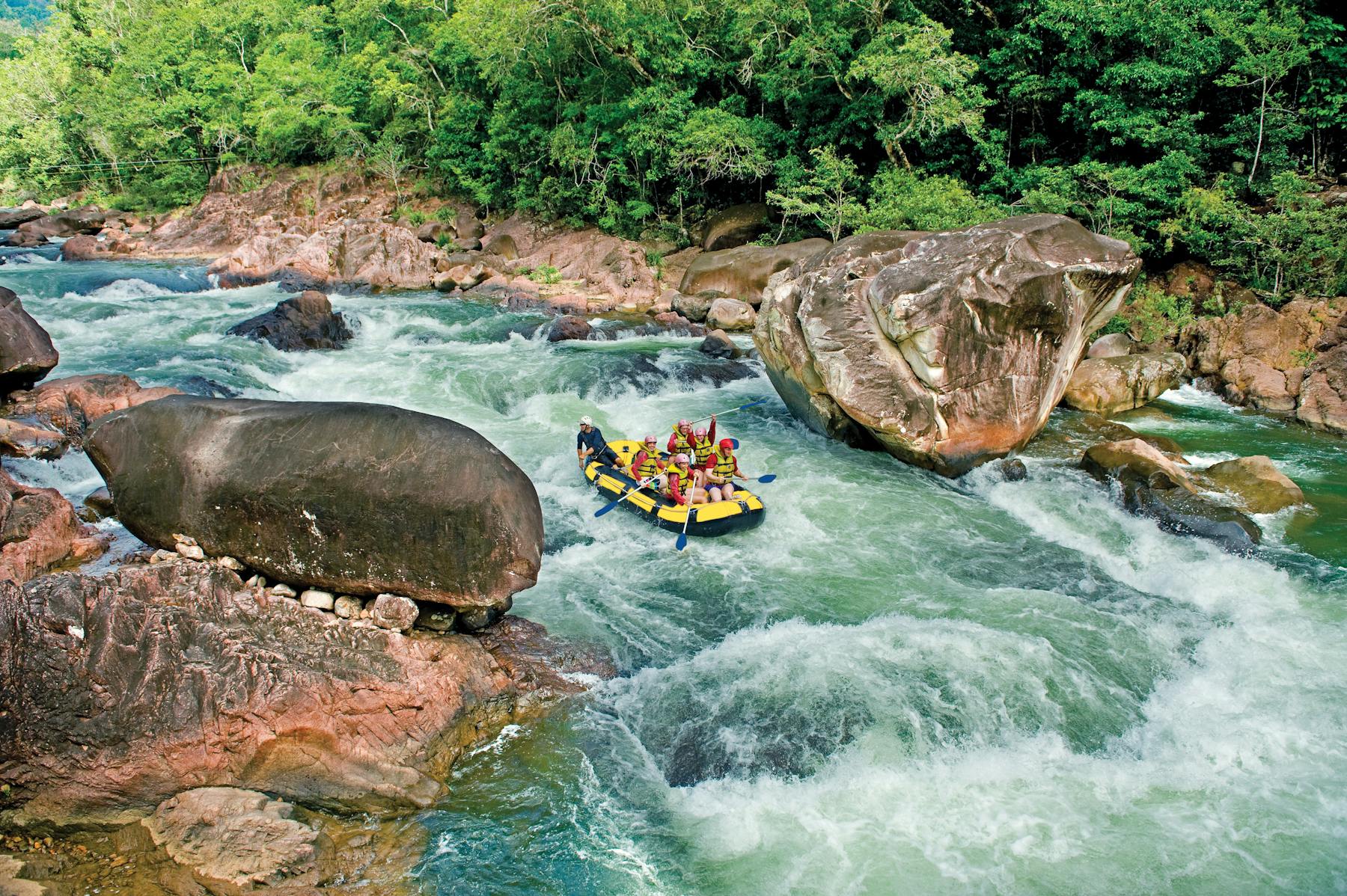 Wide shot of a rafting boat surrounded by blue water, lush rainforest and large boulders in Tully