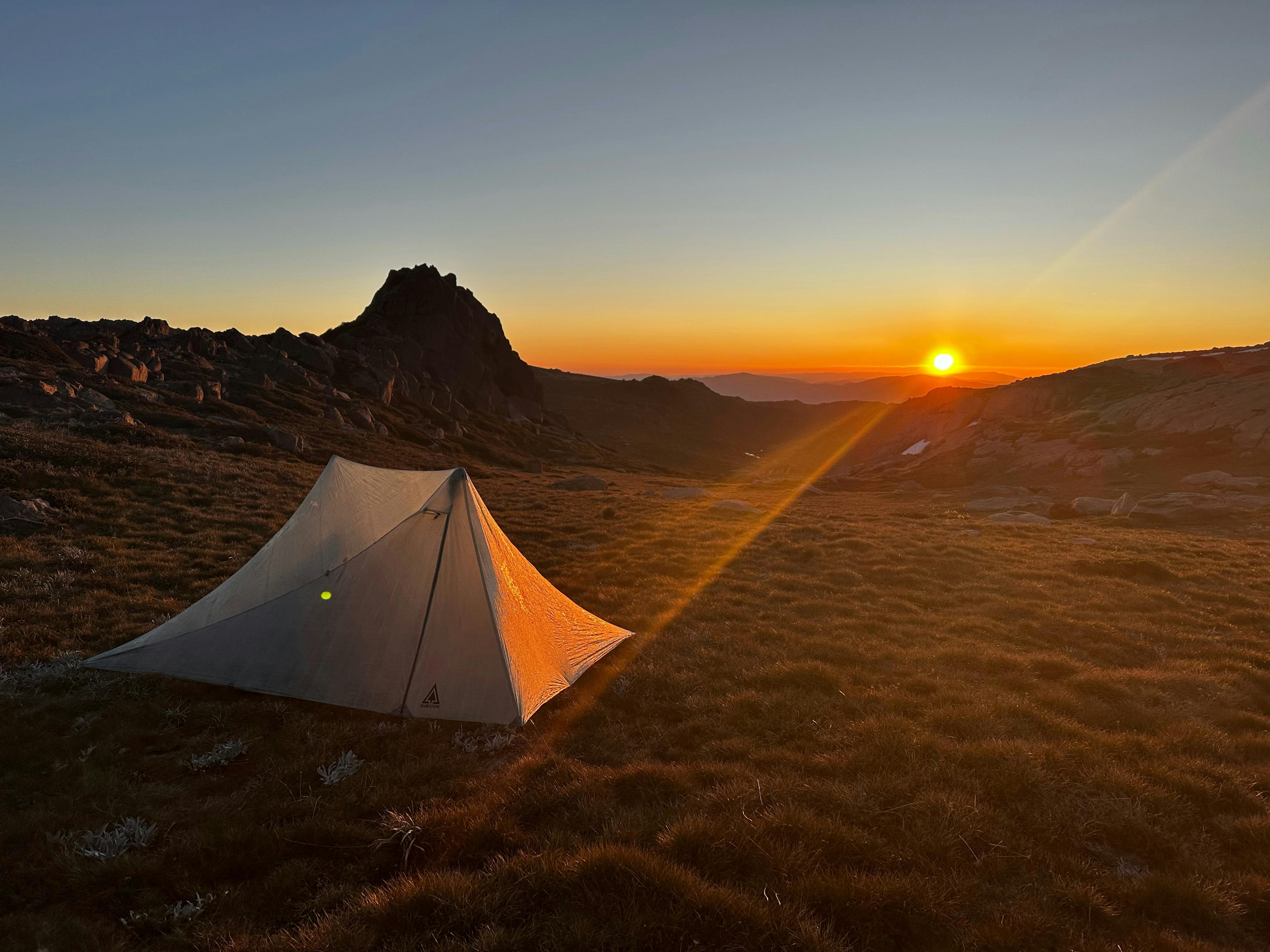 A tent on the mountains with the sun setting behind it.