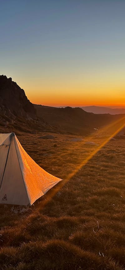 A tent on the mountains with the sun setting behind it.