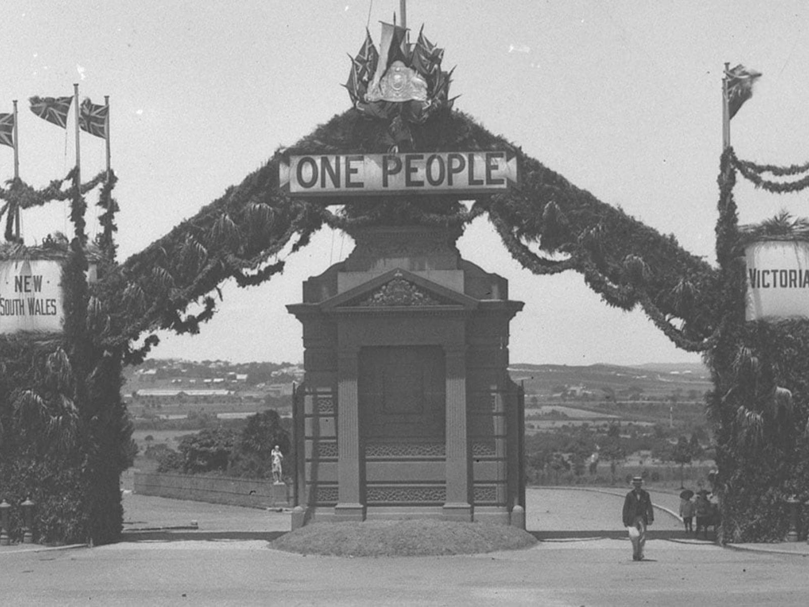Black and white photo of sandstone gates with a sign that says One People. Flags wave on the sides