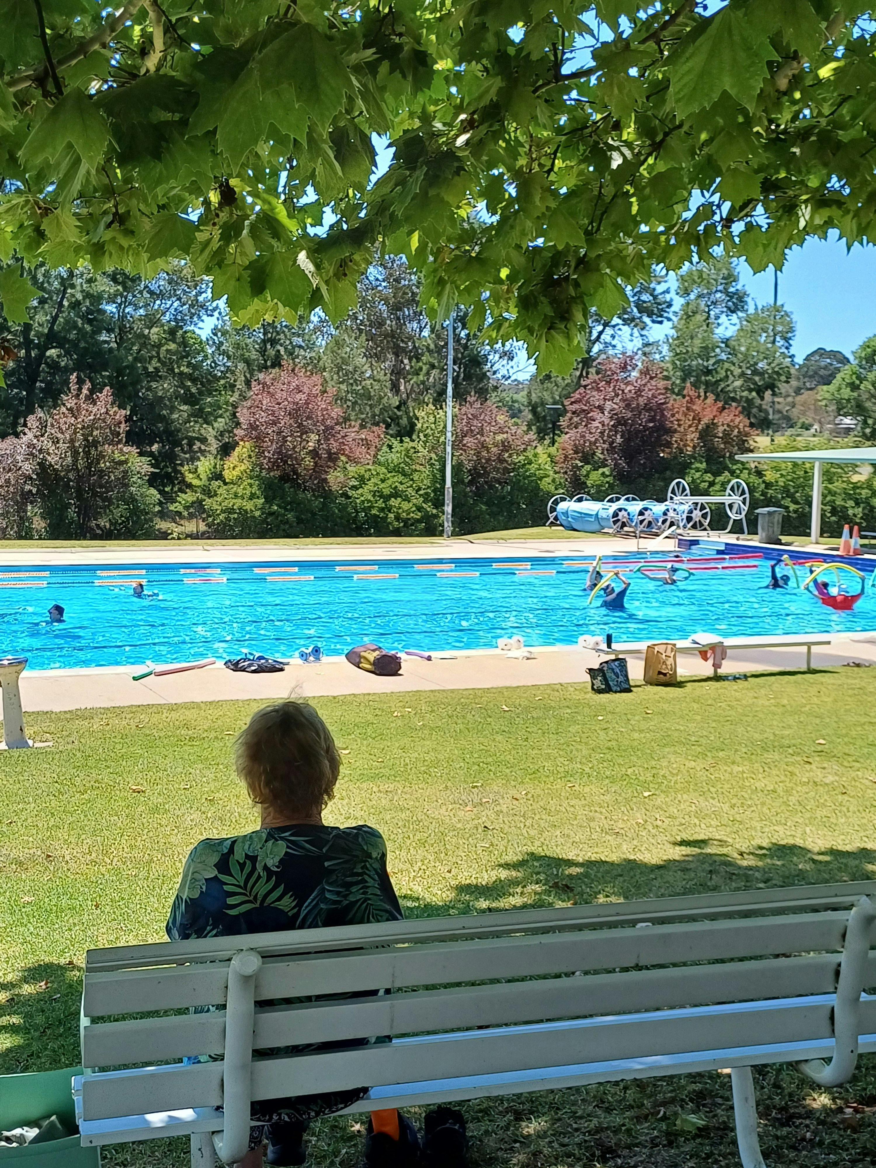 A woman sitting in the shading, watching a group of seniors doing a swimming lesson.