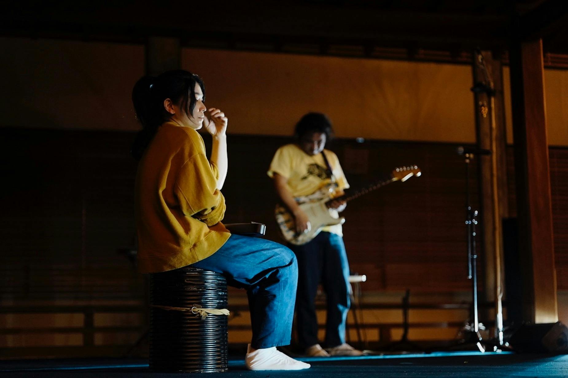 A person in the foreground sits on a keg, while a person in the background plays an electric guitar.