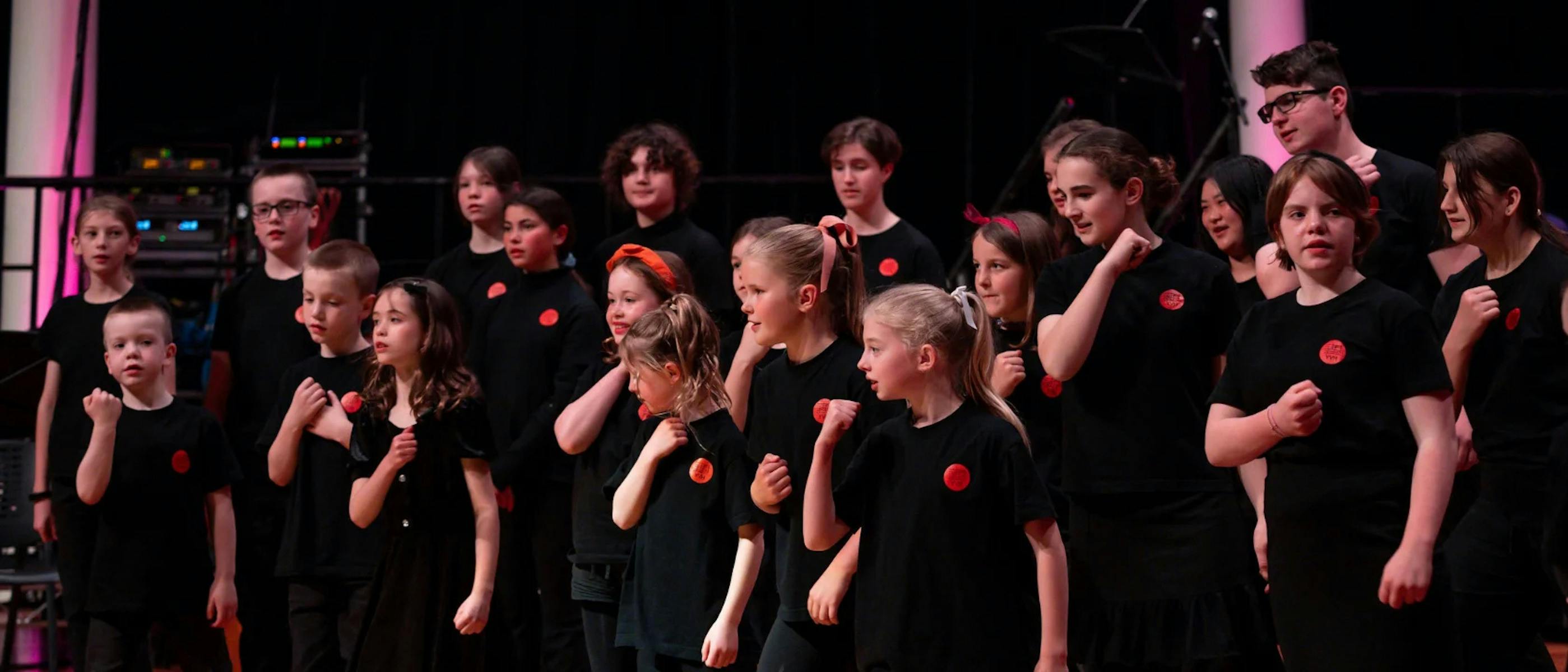 A group of children wearing black tops with red badges stand on a stage