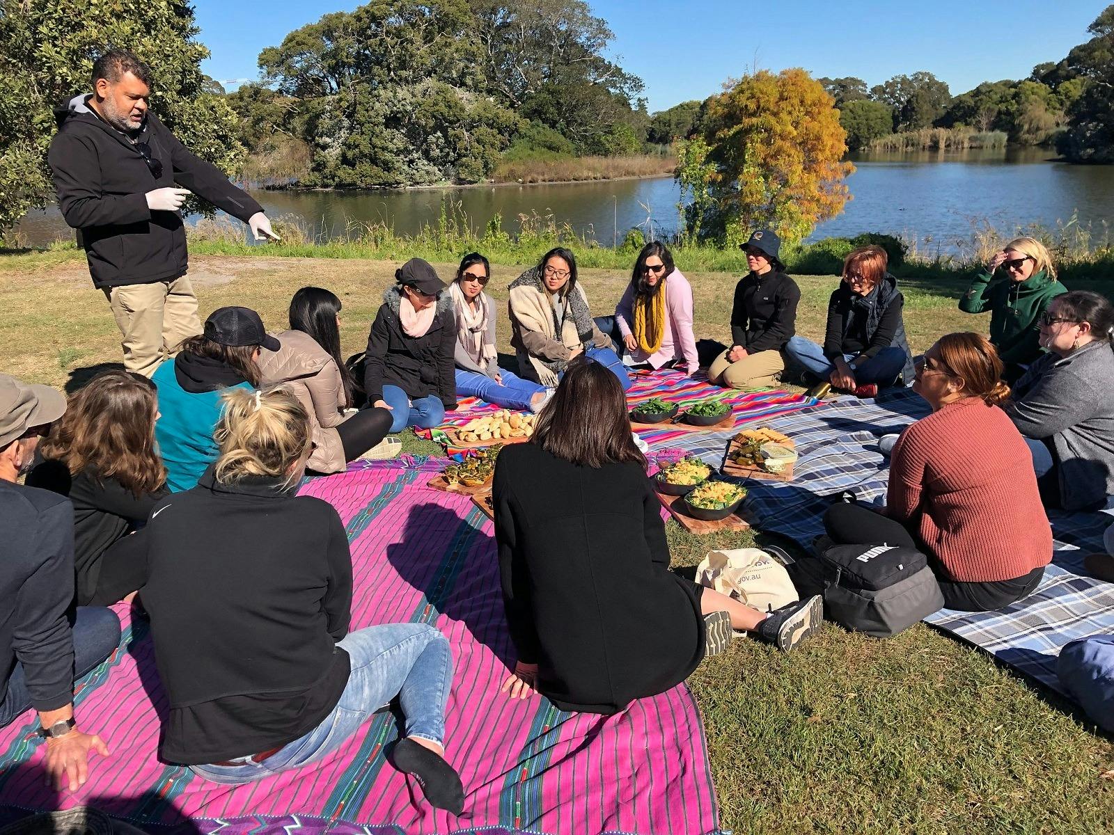 A group sitting on picnic blankets and near a lake and enjoying a morning tea