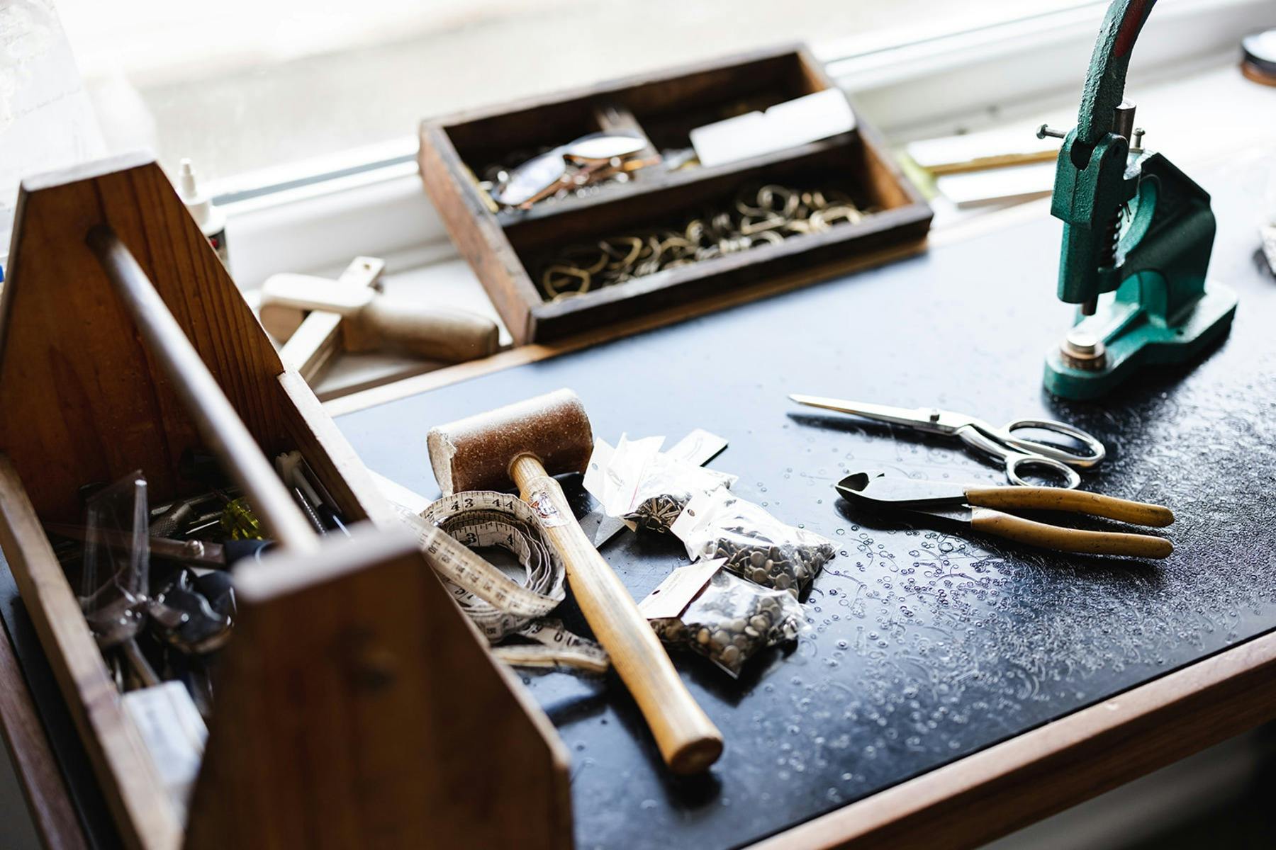 Leather crafting tools on a workbench, including a hammer, scissors, pliers, and metal hardware