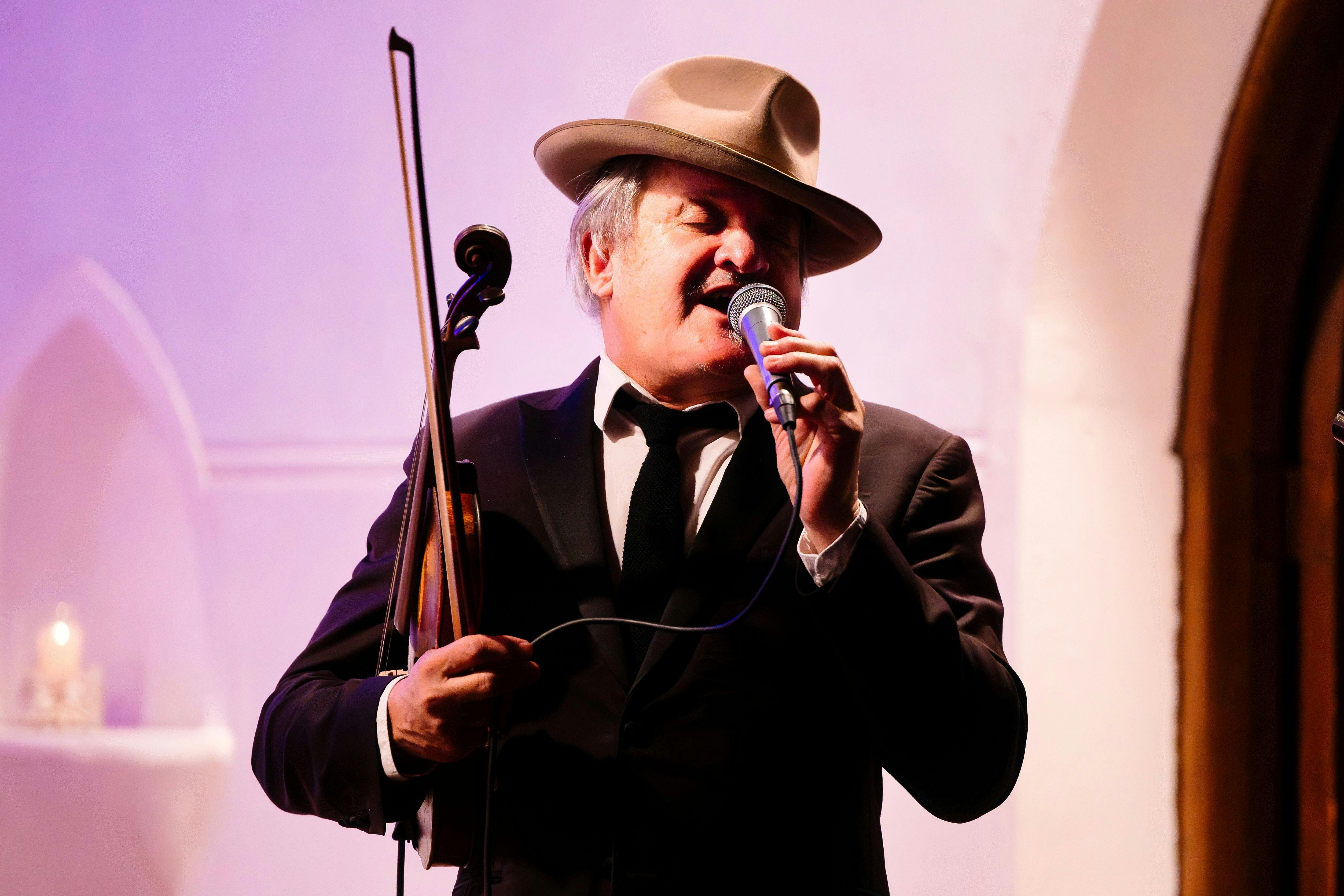 A man wearing a fedora sings into a microphone. The background lighting is pale pink and mauve